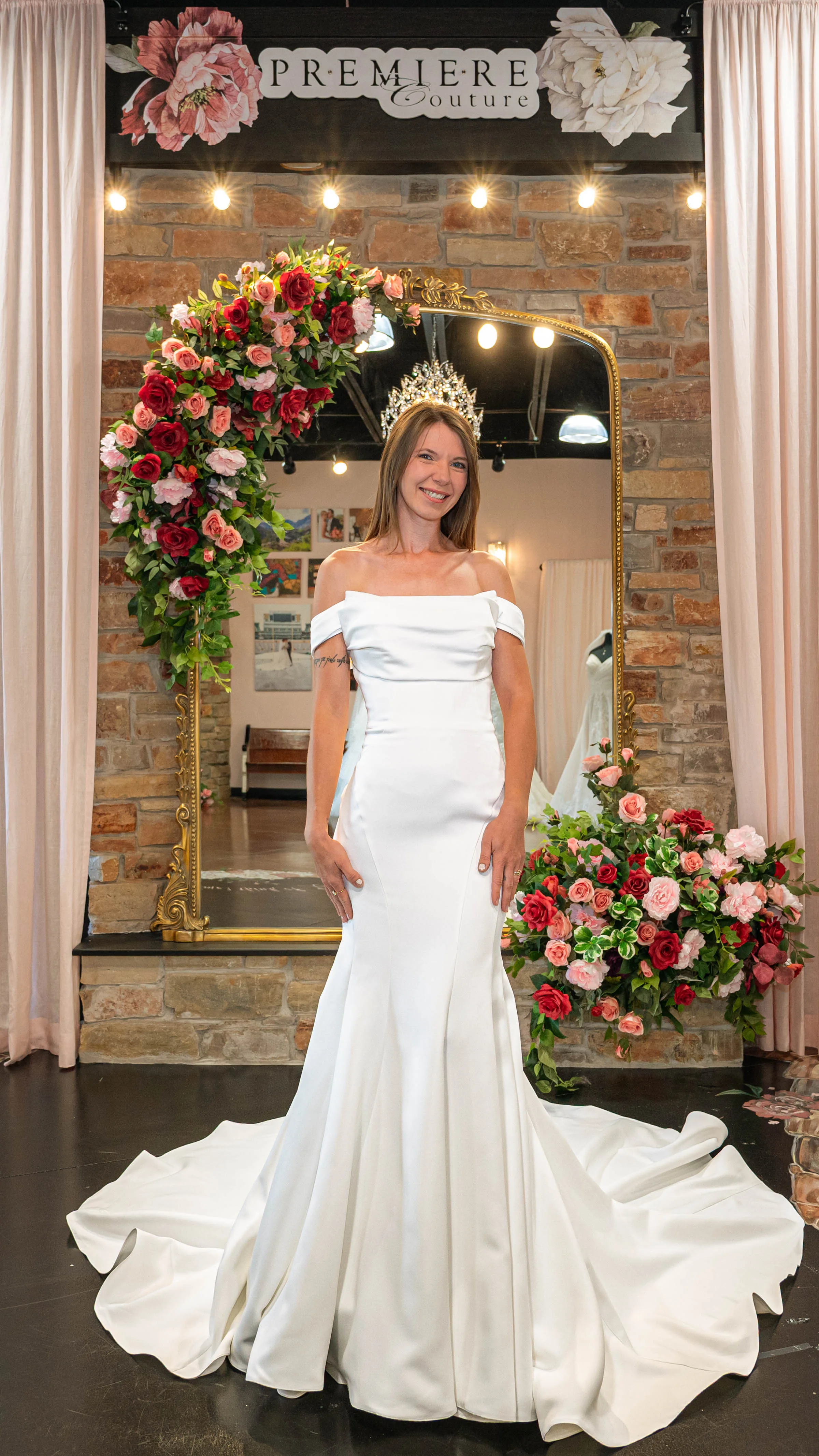 A smiling bride-to-be poses at Premiere Couture in Cambridge, Wisconsin, wearing a minimalist, off-the-shoulder fit-and-flare wedding gown with a structured satin bodice and dramatic train. She is accessorized with a sparkling bridal crown tiara, standing in front of a stone wall accented by a gold mirror draped in red and pink roses.