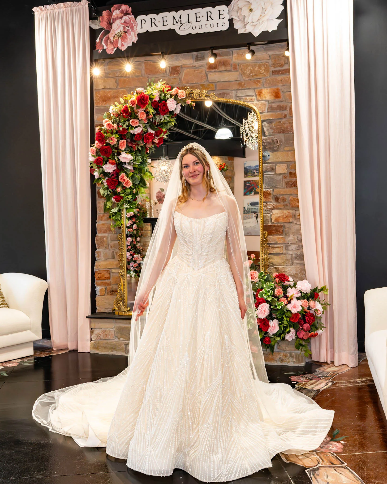 A smiling bride-to-be poses at Premiere Couture in Cambridge, Wisconsin, wearing a glamorous, heavily embellished A-line ballgown wedding dress with a strapless corset-style bodice and linear beading. She is accessorized with a sparkling tiara headband and a long, sheer tulle veil, standing in front of the boutique's signature stone wall and rose-draped gold mirror.