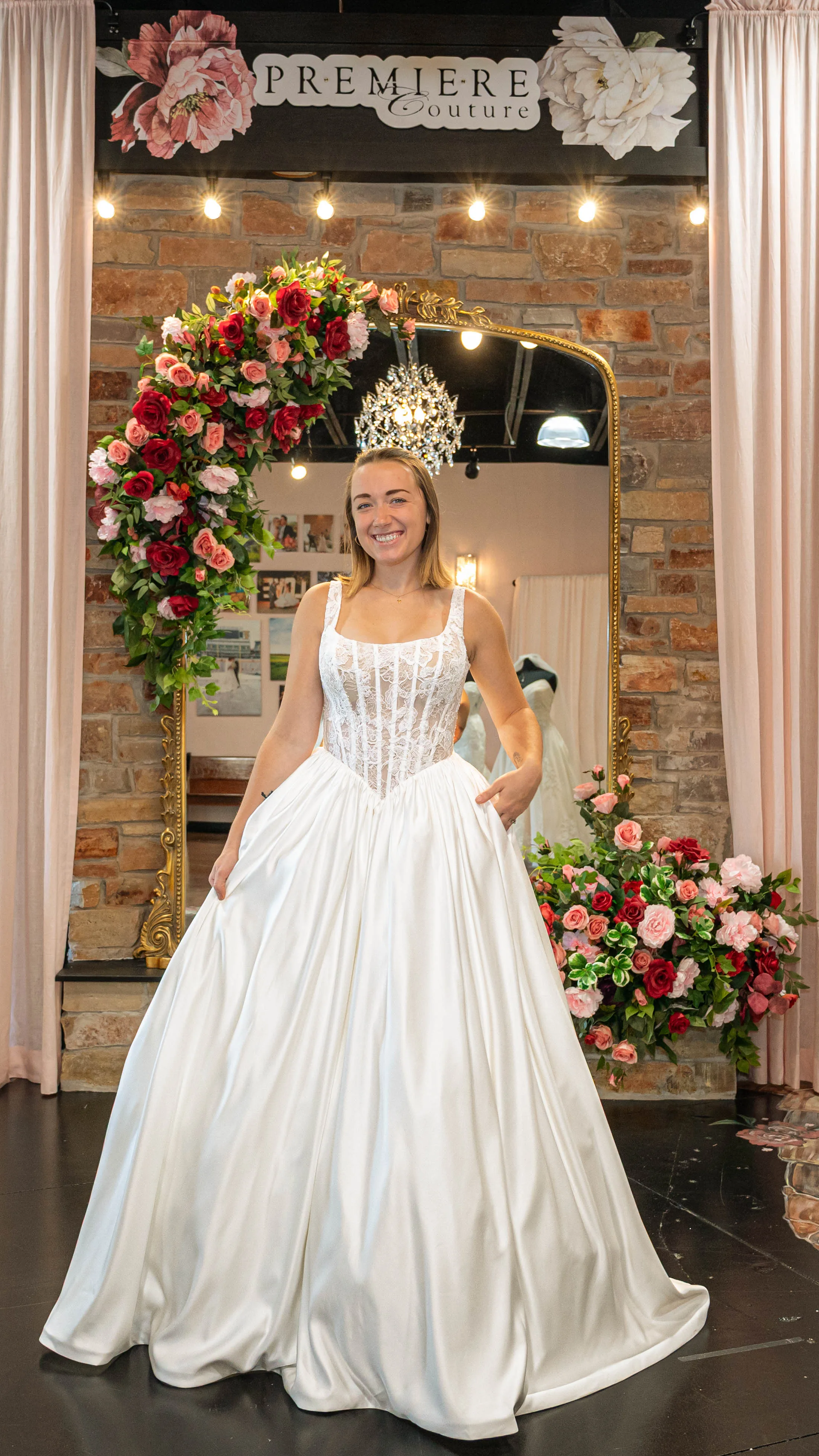 A smiling bride-to-be poses at Premiere Couture in Cambridge, Wisconsin, wearing a romantic ballgown wedding dress featuring a structured, sheer lace corset bodice with visible boning, thick shoulder straps, and a full satin skirt with pockets. She stands in front of the boutique's signature stone wall and rose-draped gold mirror.