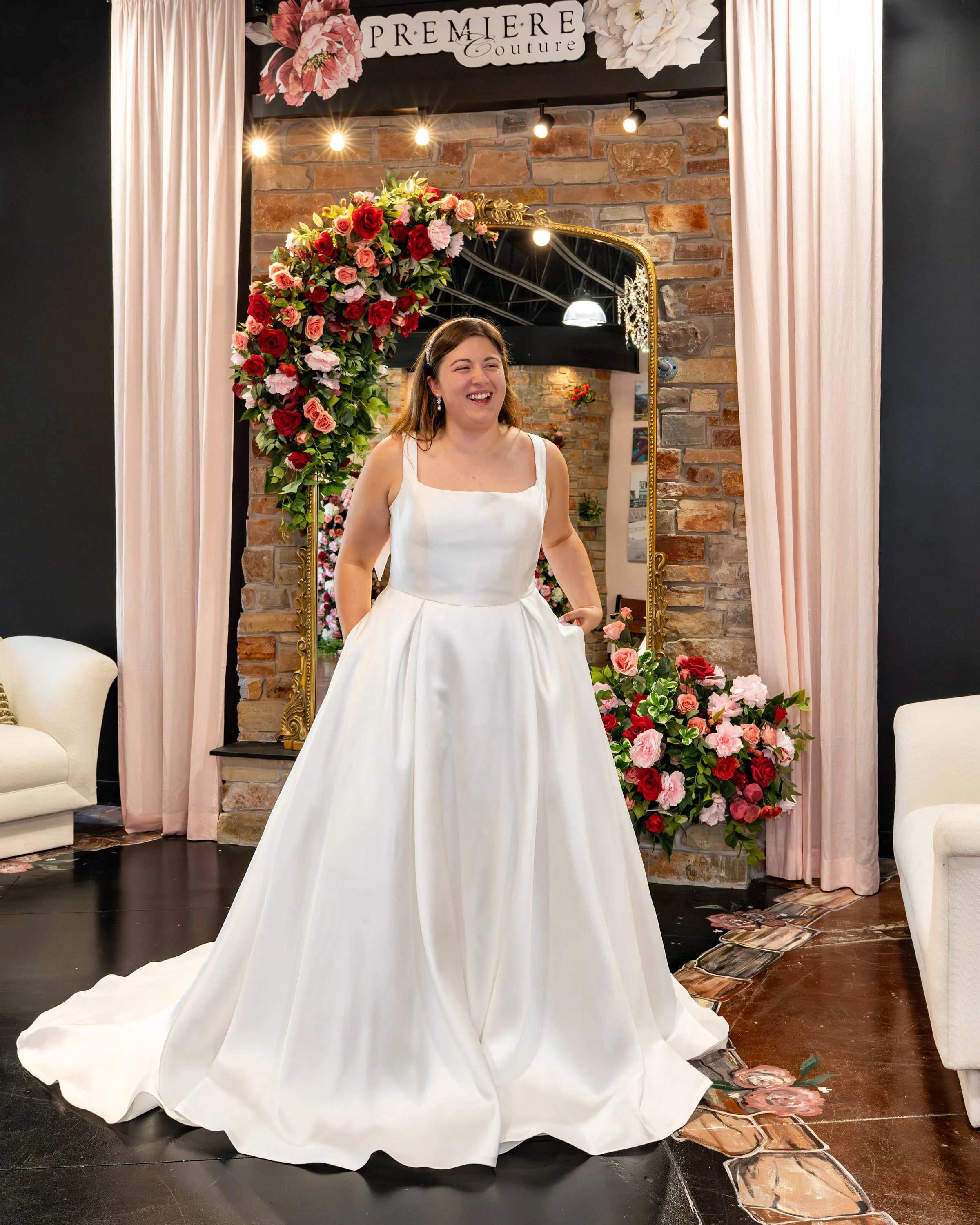 A laughing bride-to-be poses at Premiere Couture in Cambridge, Wisconsin, wearing a classic, minimalist ballgown wedding dress made of smooth satin, featuring a square neckline, wide shoulder straps, a structured bodice, and a voluminous pleated skirt with pockets. She stands in front of the boutique's signature stone wall and rose-draped gold mirror.