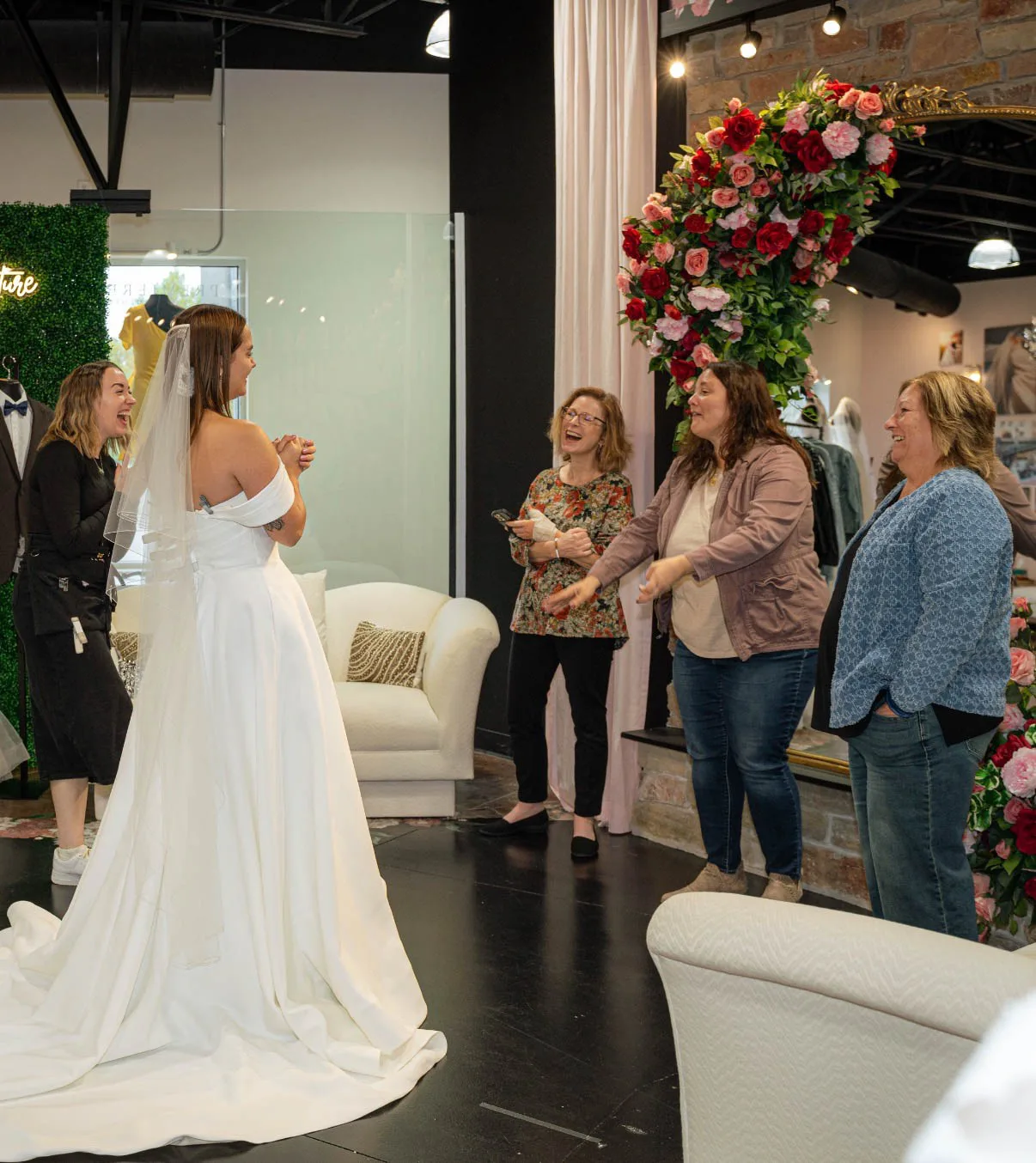 A joyful candid moment at Premiere Couture in Cambridge, Wisconsin, showing a bride from behind in an off-the-shoulder A-line wedding dress and lace-trimmed veil as her family and boutique stylist clap, laugh, and celebrate her finding her dress.