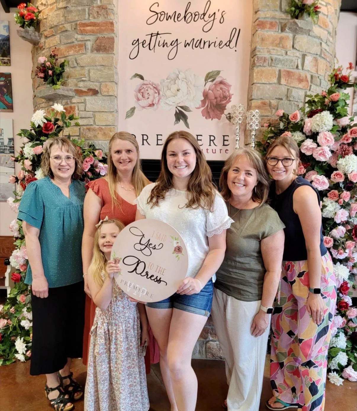 A happy bride-to-be stands with her group of friends and family at Premiere Couture in Cambridge, Wisconsin, holding a round floral sign that reads "I Said Yes to the Dress." They are posing in front of a stone wall backdrop featuring floral arrangements and a sign that says "Somebody's getting married!"