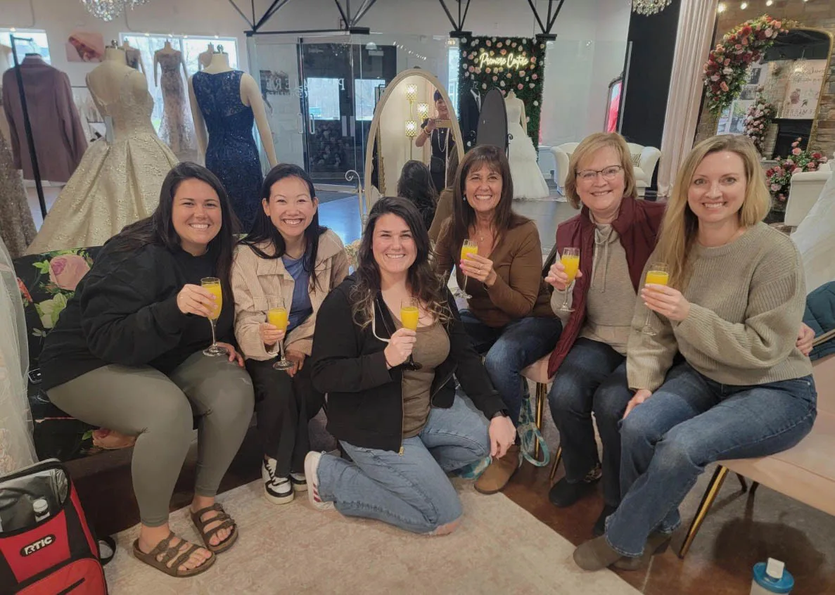 Six smiling women sit together and toast with glasses of champagne or mimosas during a bridal appointment at Premiere Couture in Cambridge, Wisconsin. Gowns on mannequins, a mirror, and the boutique showroom are visible in the background.