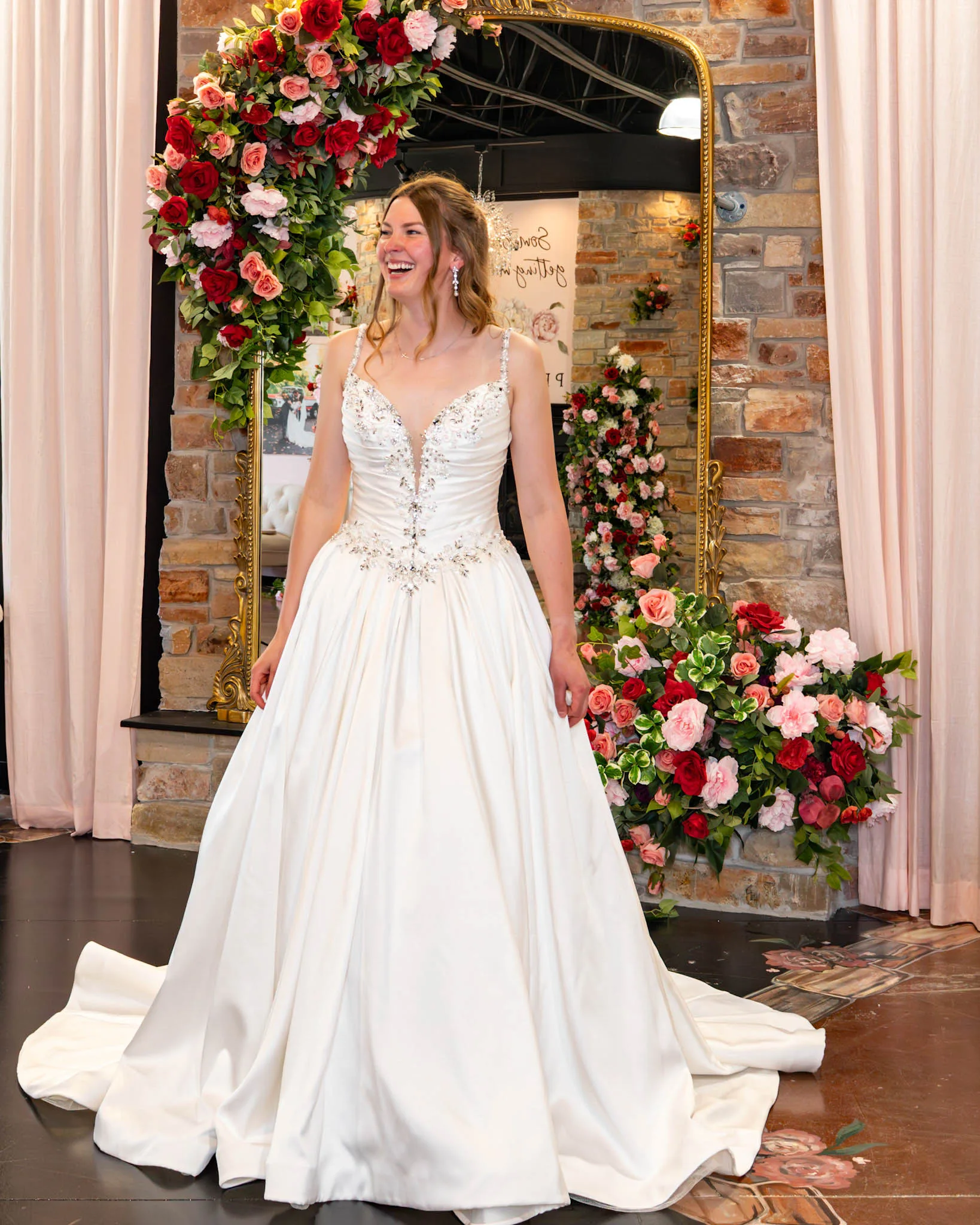 A laughing bride-to-be poses at Premiere Couture in Cambridge, Wisconsin, wearing a glamorous A-line satin ballgown wedding dress featuring a ruched V-neck bodice, delicate beaded straps, and intricate crystal embellishments along the plunge neckline and drop waist. She stands in front of the boutique's signature stone wall and rose-draped gold mirror.