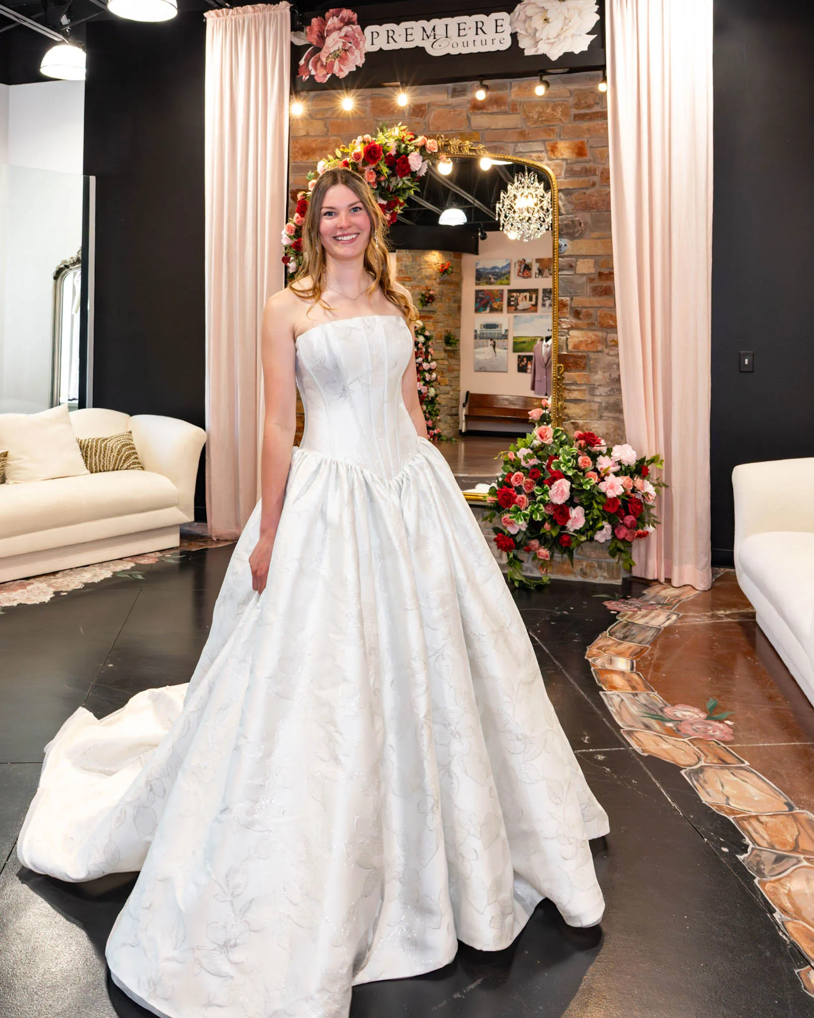 A smiling bride-to-be poses at Premiere Couture in Cambridge, Wisconsin, wearing a romantic jacquard or brocade ballgown wedding dress featuring a subtle metallic floral pattern, a strapless corset-style bodice with visible structure, a dropped basque waistline, and a full pleated skirt. She stands in front of the boutique's signature stone wall and rose-draped gold mirror.
