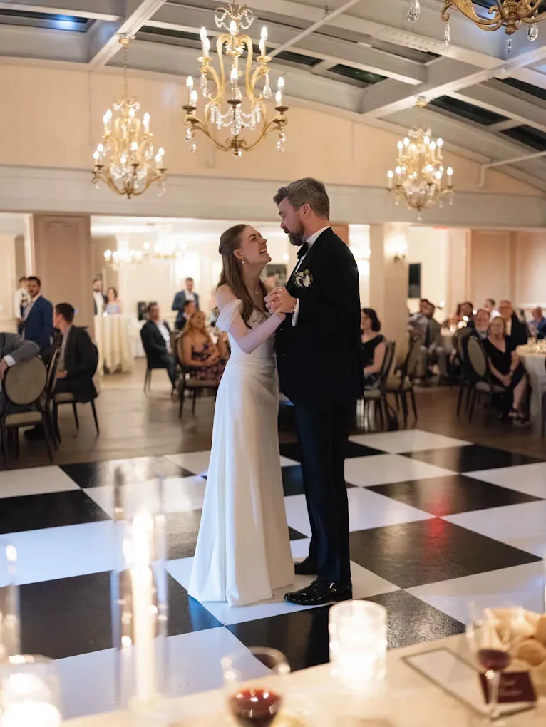 A bright, elegant photograph capturing a bride and groom laughing together during their first dance on a classic black and white checkered dance floor. The bride wears a flowing, off-the-shoulder white wedding gown, and the groom wears a black tuxedo. They are surrounded by seated wedding guests under warm, glowing crystal chandeliers at a reception entertainment setup managed by Juke Box Bandstand.