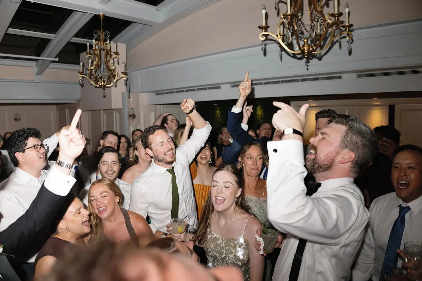 A high-energy, crowded wedding dance floor filled with smiling guests singing along with their hands in the air during a reception entertainment experience by Juke Box Bandstand in Madison, Wisconsin. The groom, in a white shirt and suspenders, sings passionately alongside the bride in a shimmering dress, surrounded by friends holding drinks under bright venue lighting and ornate gold chandeliers.