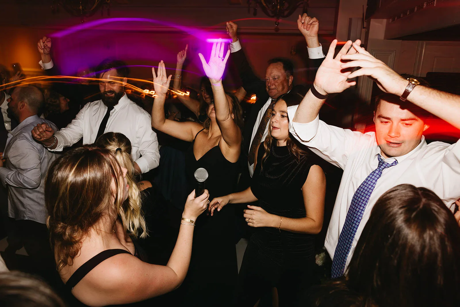 A lively, energetic photograph of a packed wedding dance floor packed with guests dancing with their hands in the air, entertained by Juke Box Bandstand in Madison, Wisconsin. In the foreground, a woman sings enthusiastically into a wireless microphone provided by the DJ, surrounded by friends in formal attire under vibrant purple and red event party lighting.