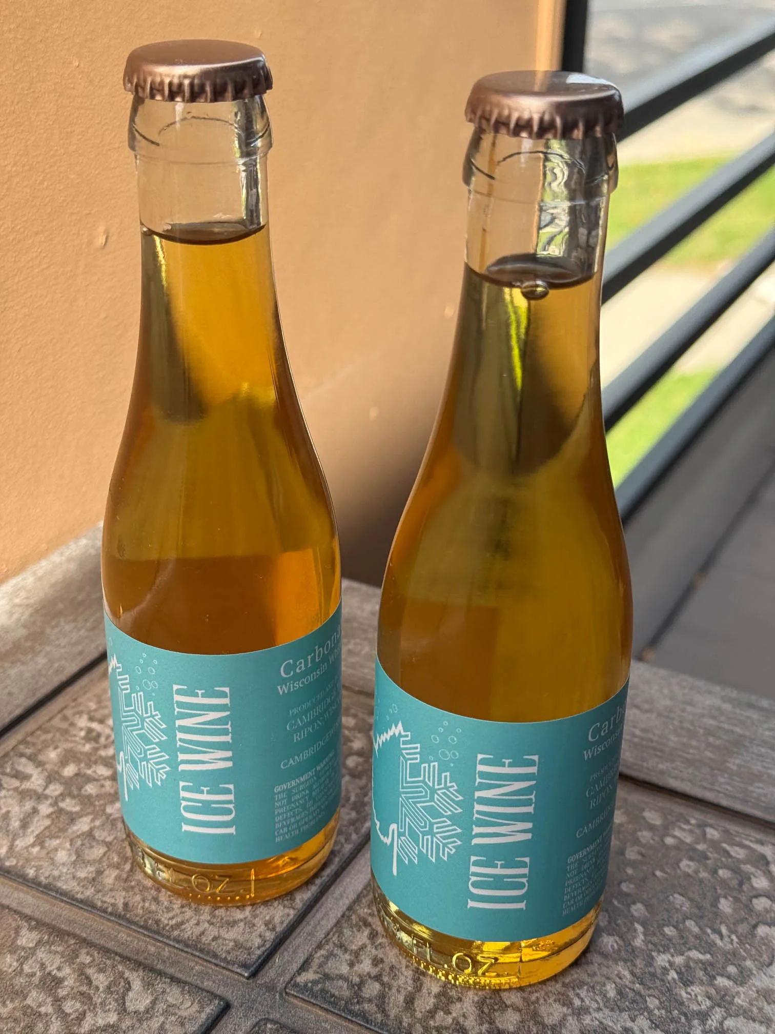 Two bottles of carbonated Ice Wine by Cambridge Winery with a light blue snowflake label sitting outside on a textured ledge.