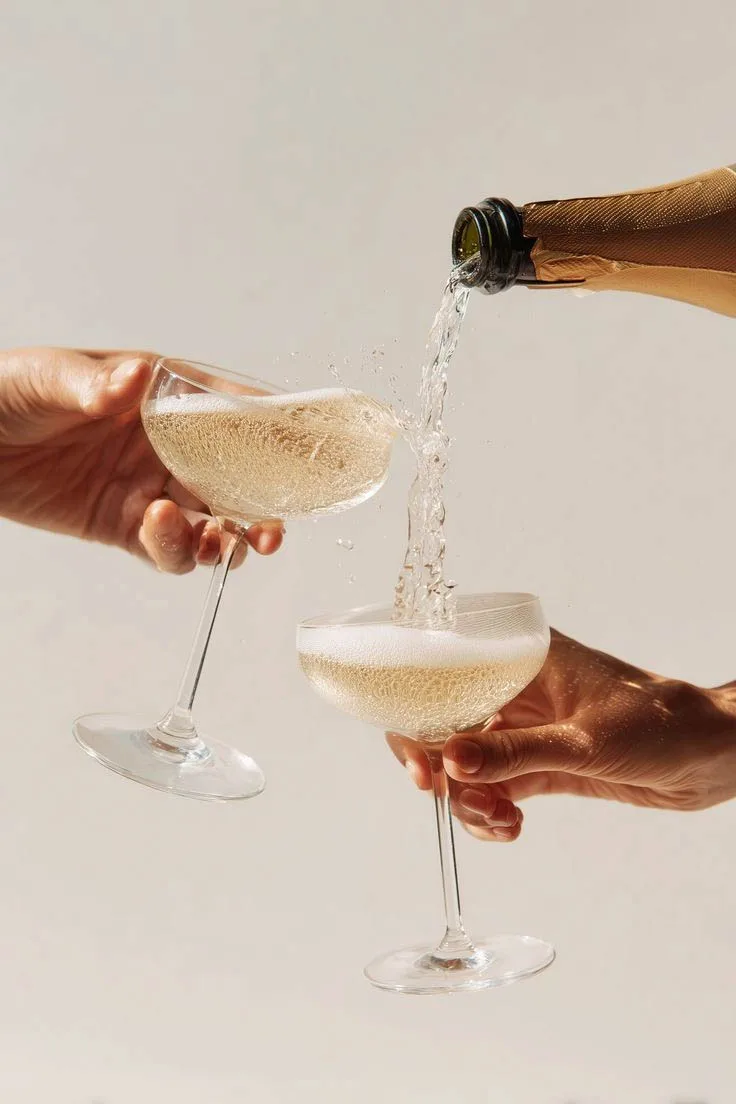 Sparkling wine is poured from a bottle into two coupe glasses held by hands, creating a splash and bubbling foam, featuring selections from Cambridge Winery.