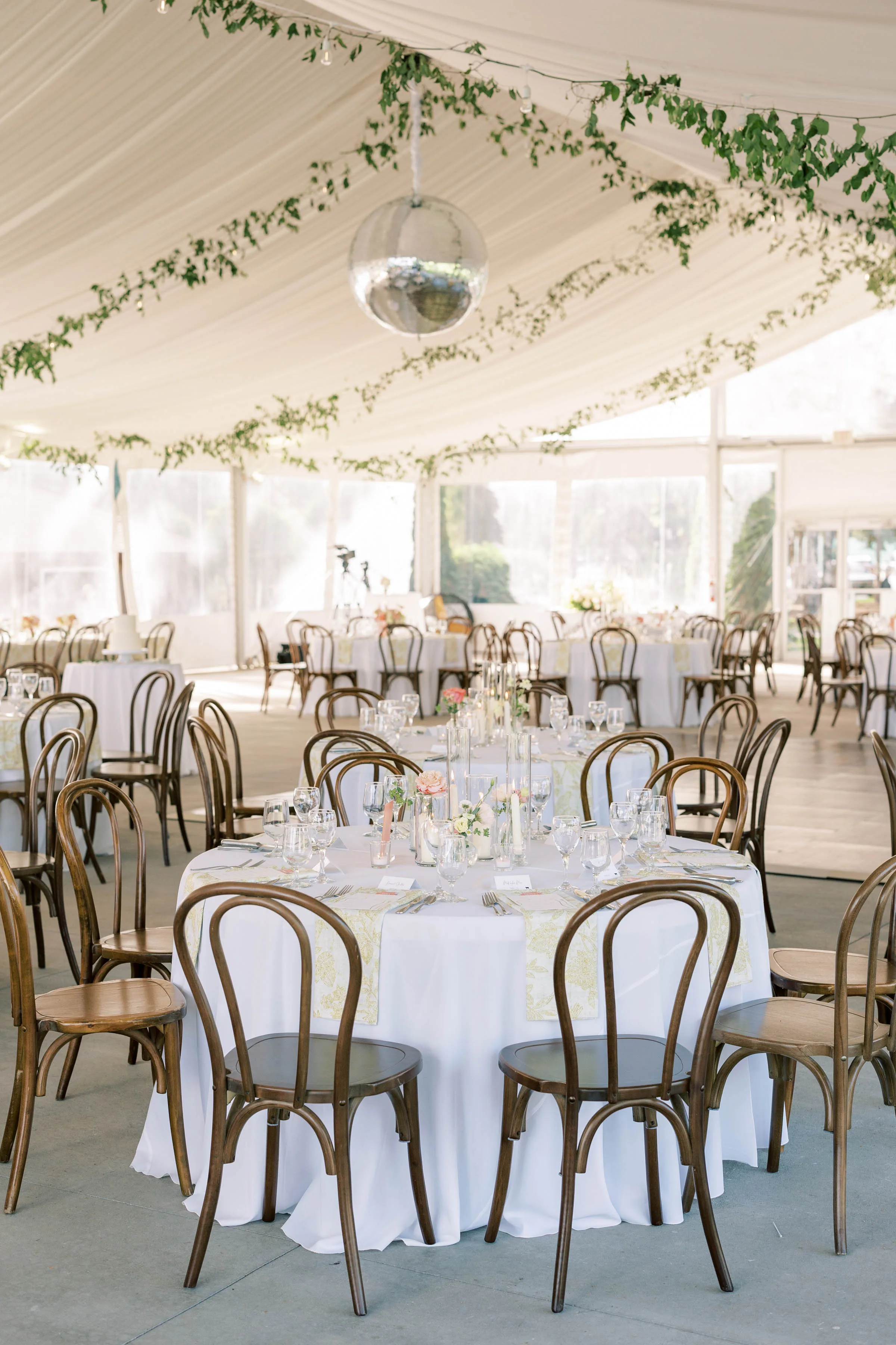 Interior of an Event Essentials wedding tent featuring round tables with white linens, wooden bentwood chairs, delicate floral centerpieces, and a large disco ball hanging from draped ceiling fabric.