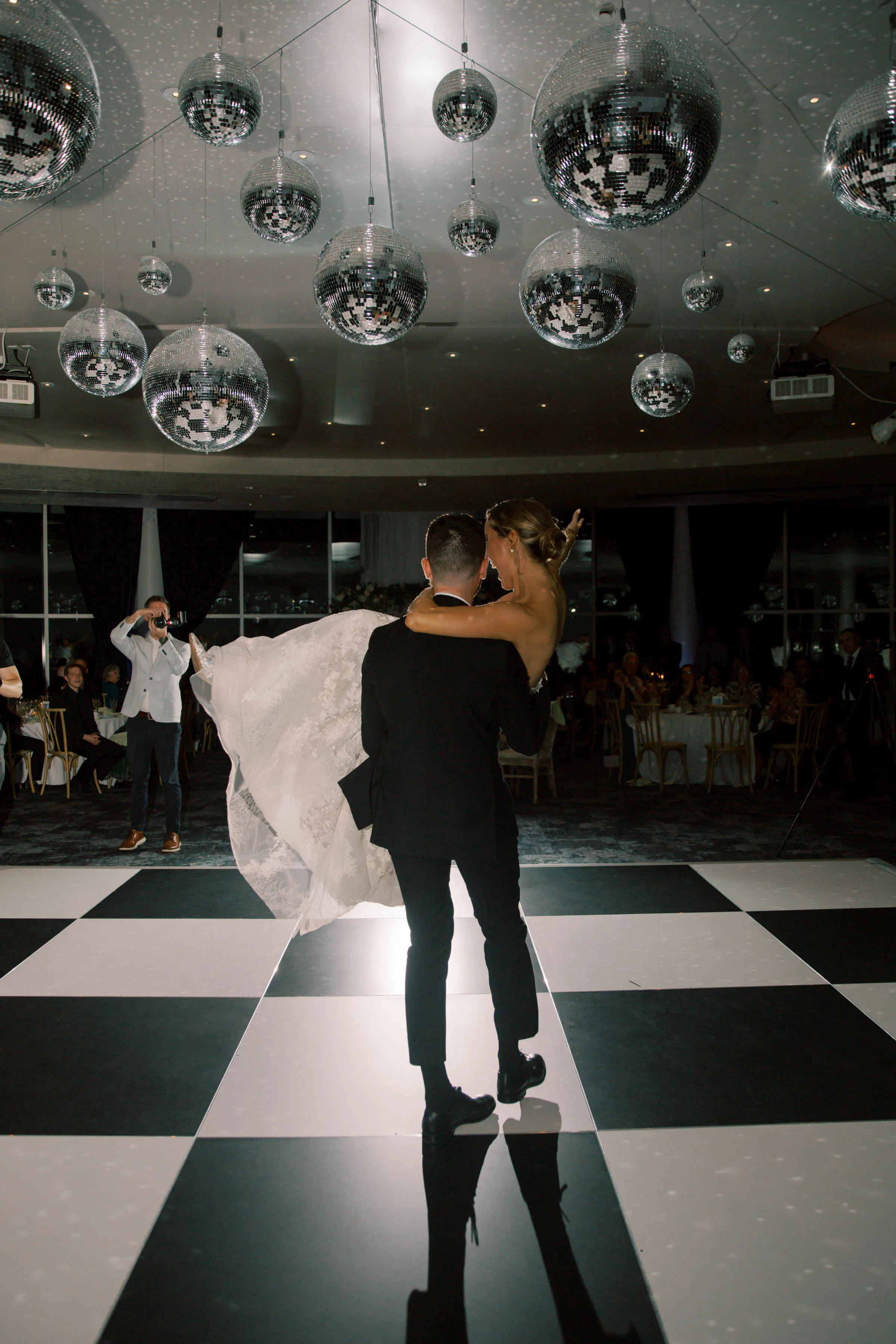 An Event Essentials black and white checkered dance floor installed for a wedding reception, featuring a groom carrying his bride under an overhead cluster of multiple hanging disco balls.