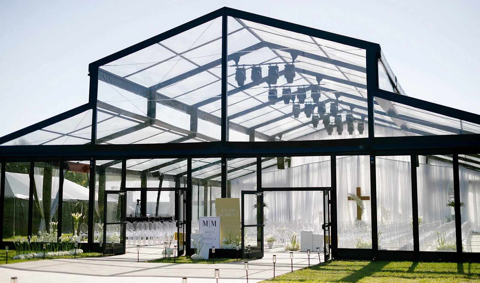 An Event Essentials clear-top structure tent with black framing and glass double doors, showcasing an upscale wedding ceremony setup inside with white drapery, professional overhead truss lighting, a large wooden cross, and rows of clear ghost chairs.