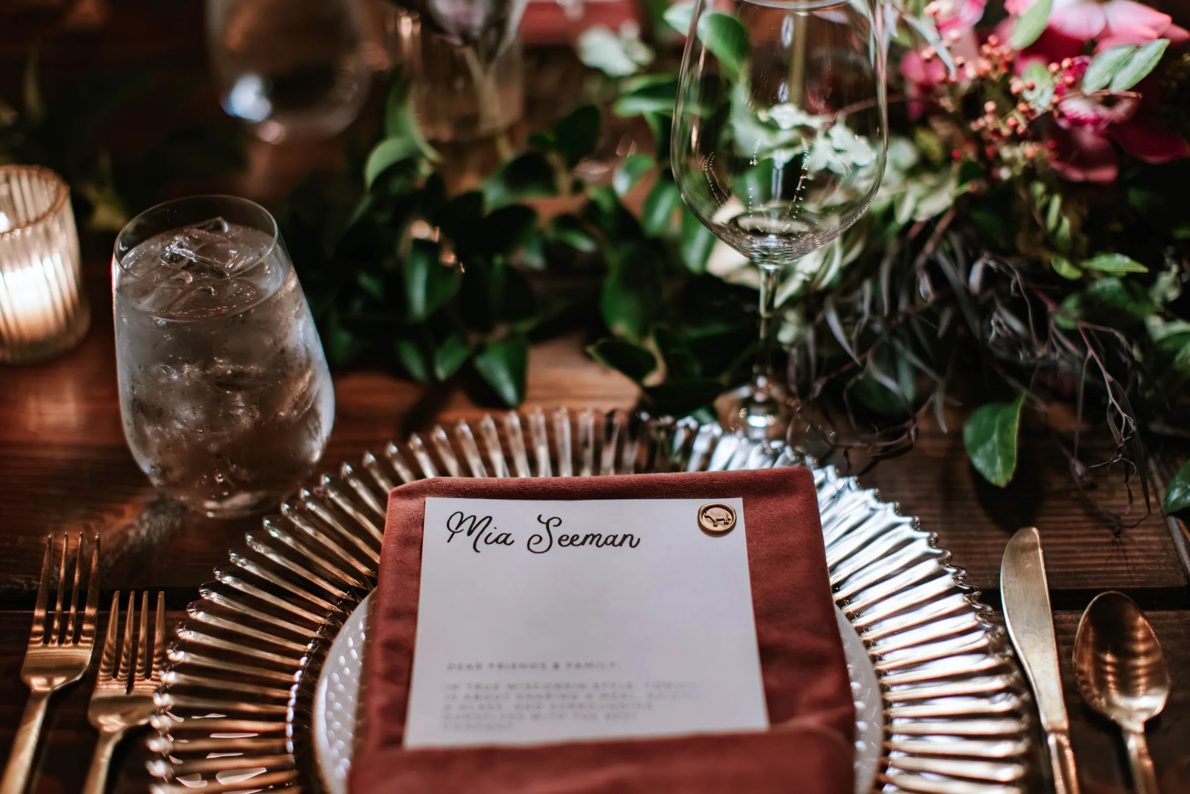 An Event Essentials table setting featuring a clear sunburst glass charger plate with a gold rim, a terracotta velvet napkin, a custom menu card, brushed gold flatware, and crystal glassware.