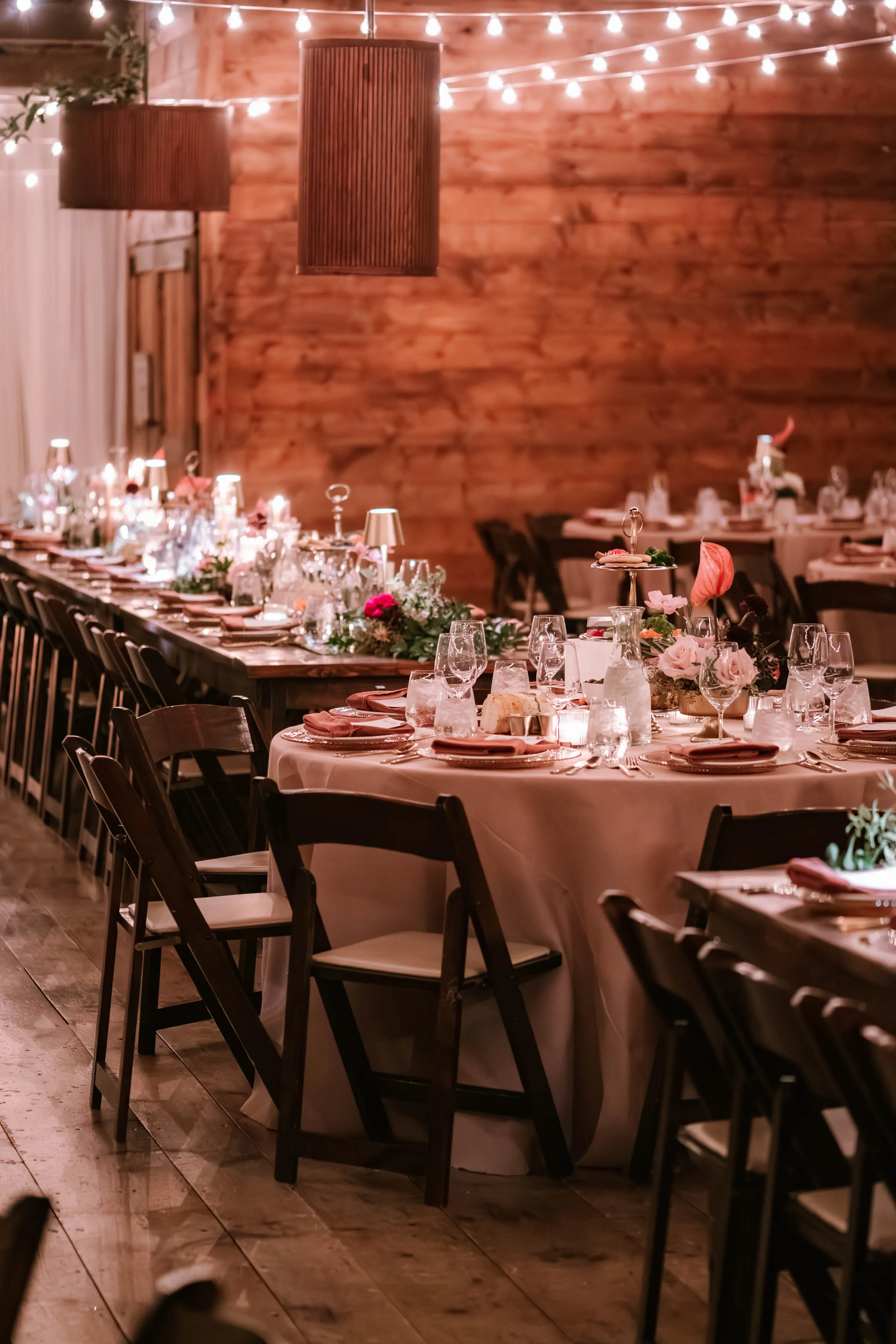 An Event Essentials barn wedding reception setup featuring wooden farm tables mixed with round tables covered in blush linens, paired with dark wood folding chairs with white cushions under warm string lighting.