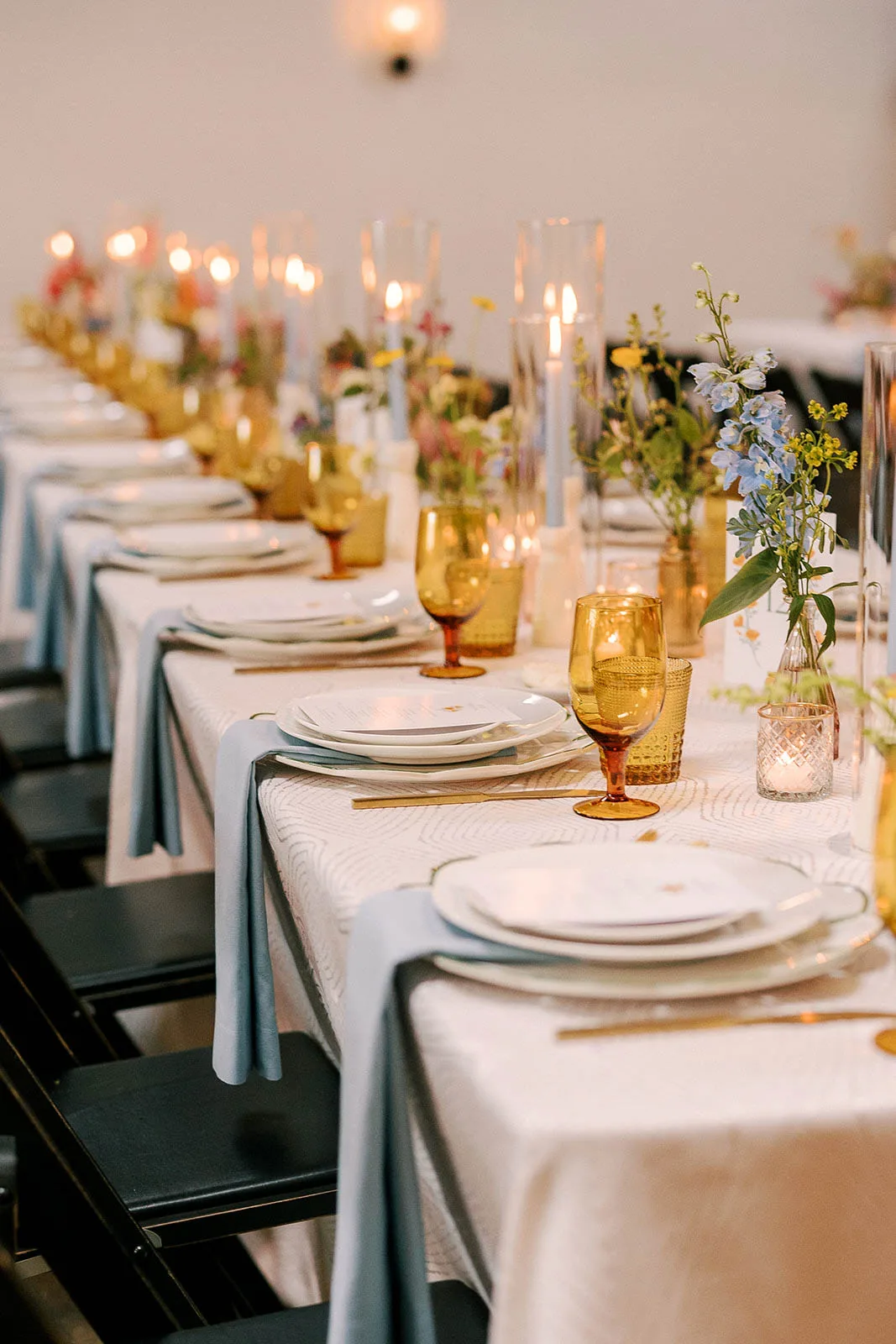 An Event Essentials table scape featuring textured white linens, dusty blue napkins draped over white dinner plates, amber glass goblets, and gold flatware flanked by glass cylinder taper candles.