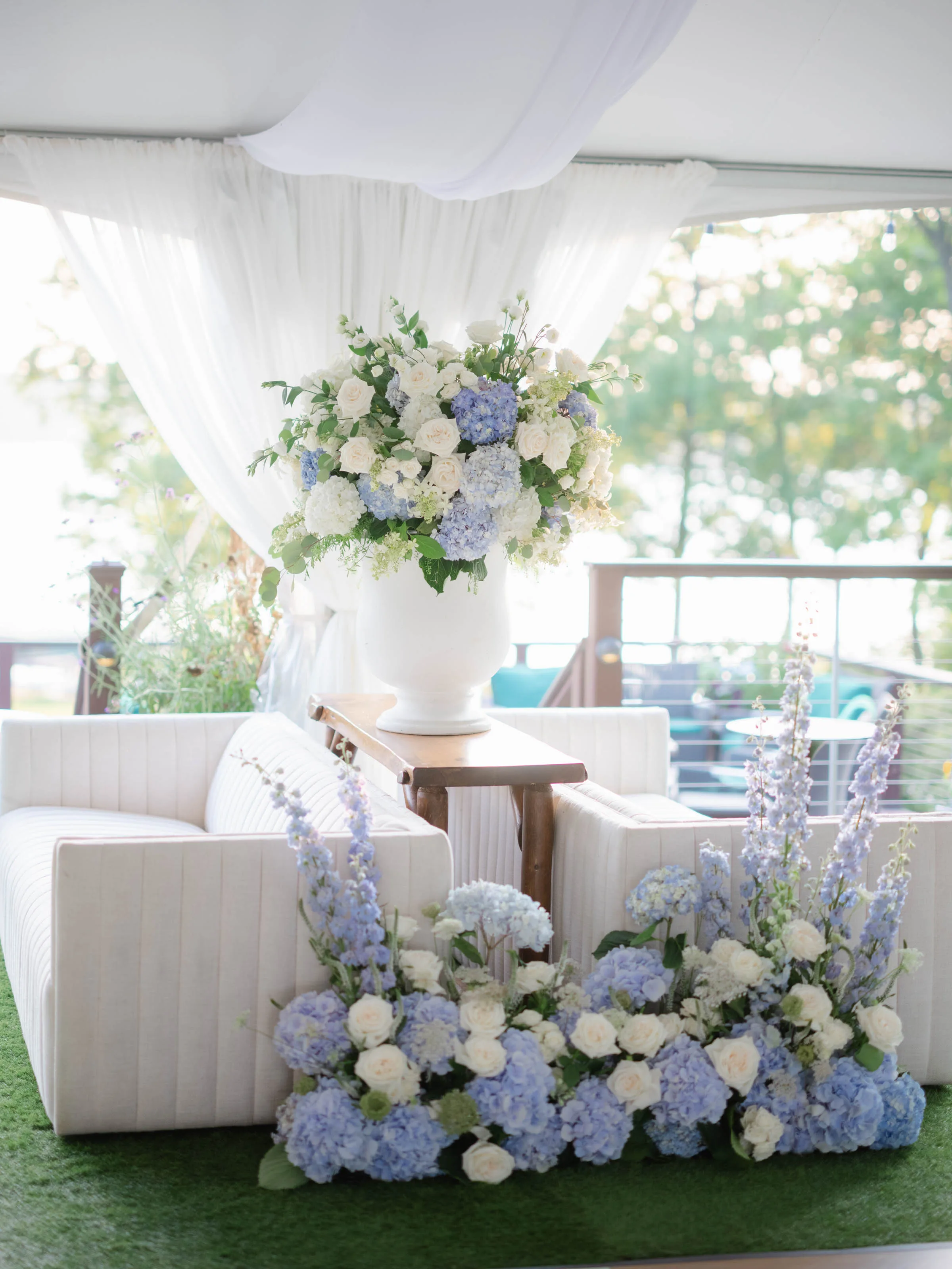 An Event Essentials outdoor event lounge featuring two white channel-tufted sofas facing each other, divided by a wooden console table with a large floral arrangement and white fabric drapery in the background.