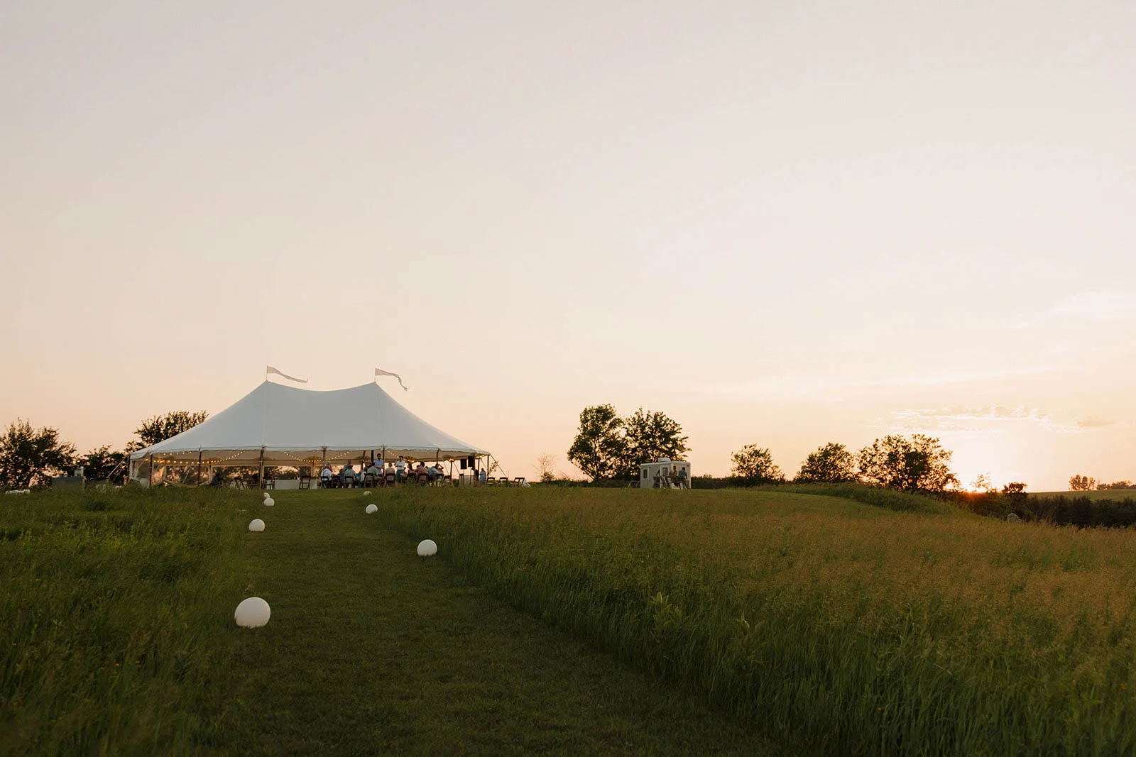 A sprawling Midwest outdoor wedding reception featuring a large Event Essentials pole tent glowing under a golden sunset sky on a rural grassy hill.