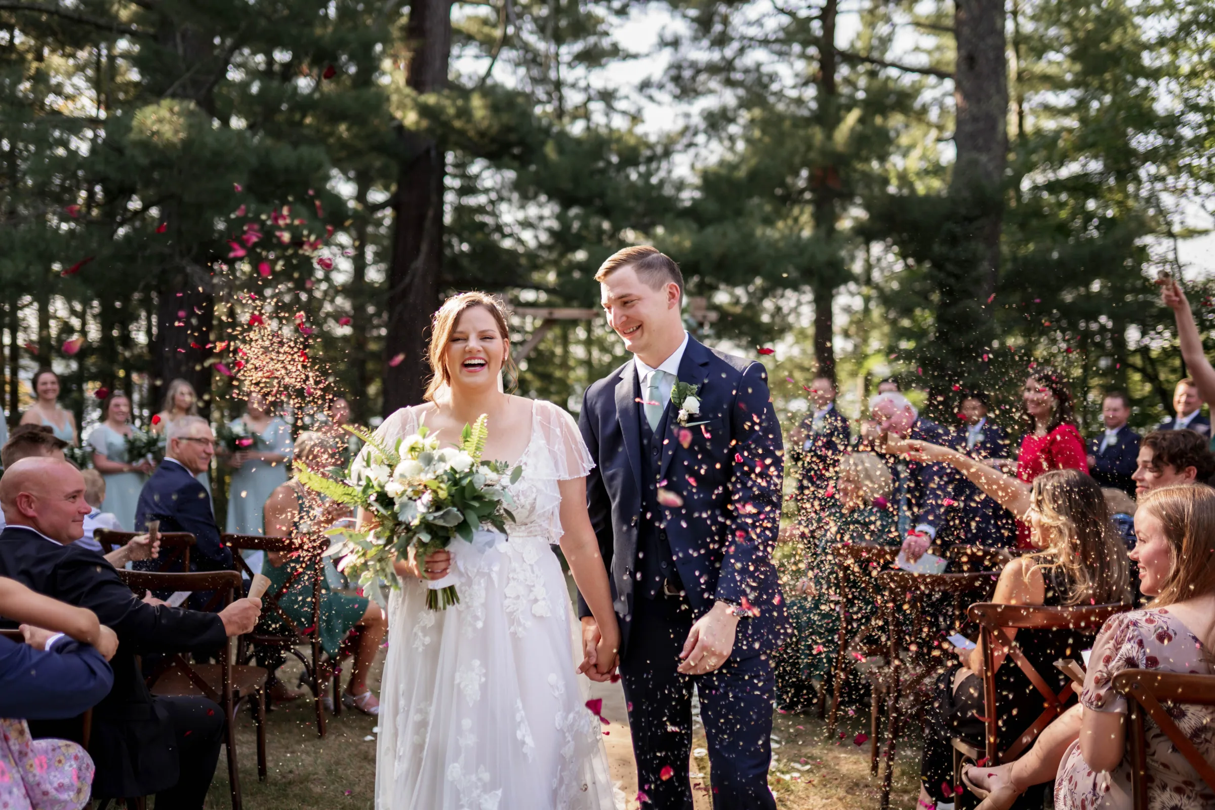 Outdoor woodland wedding photography in Madison, WI, by Blu Photography & Design LLC, featuring a smiling bride and groom walking down the aisle together as wedding guests throw colorful flower petals.