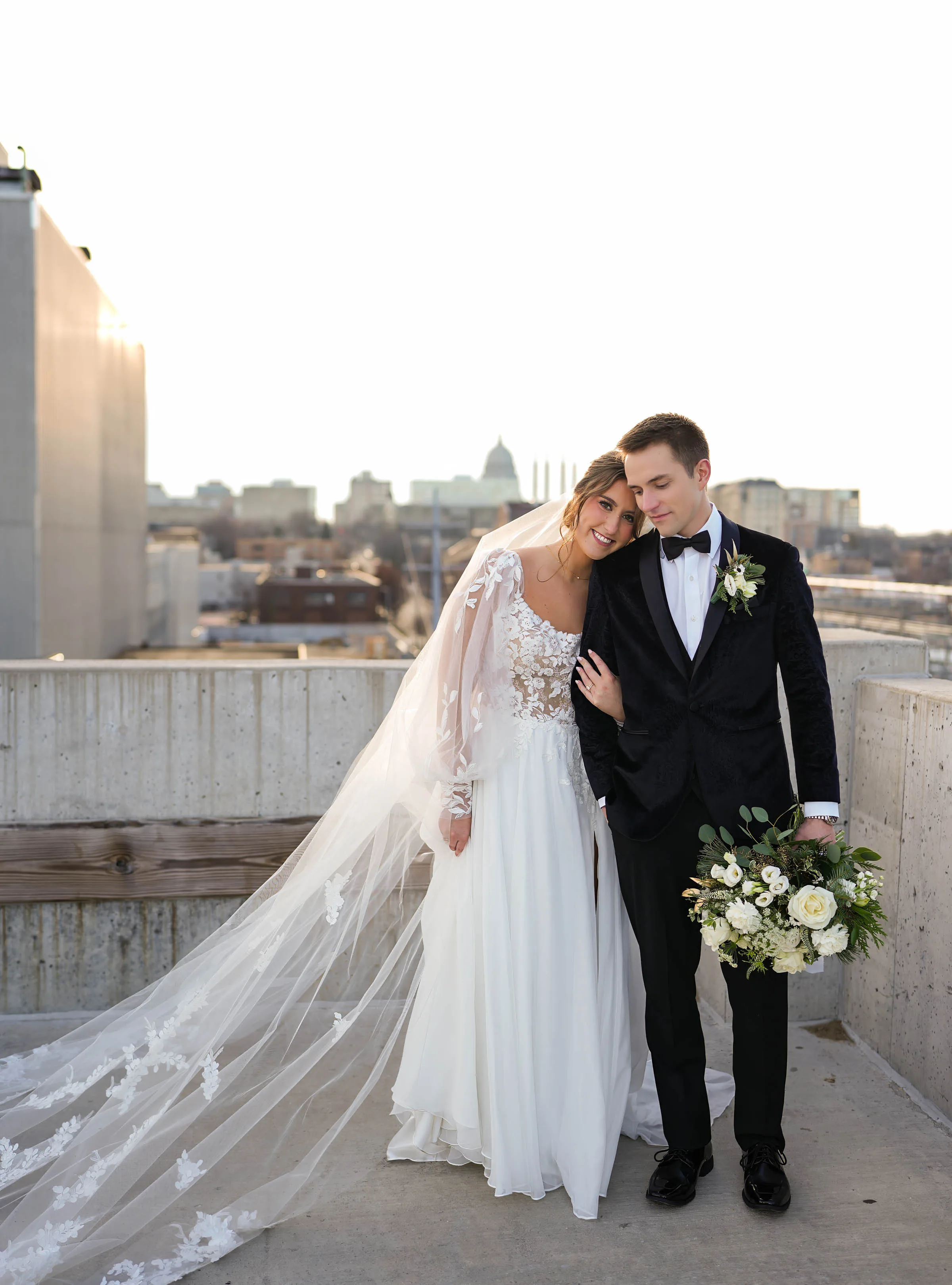 Rooftop wedding photography with the Wisconsin State Capitol skyline in Madison, WI, by Blu Photography & Design LLC, showcasing a bride in a long-sleeve gown and a groom in a black tuxedo holding a white bouquet.