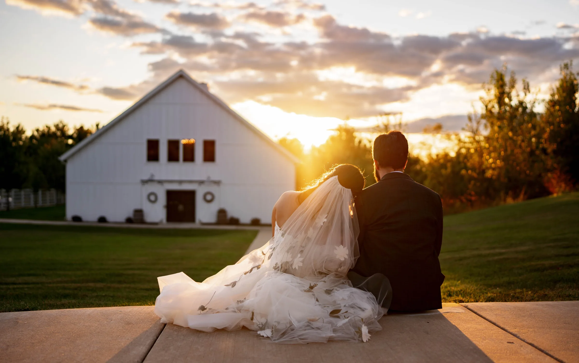 Golden hour barn wedding photography in Madison, WI, by Blu Photography & Design LLC, showing a bride and groom sitting on a concrete path looking toward a modern white barn venue at sunset.