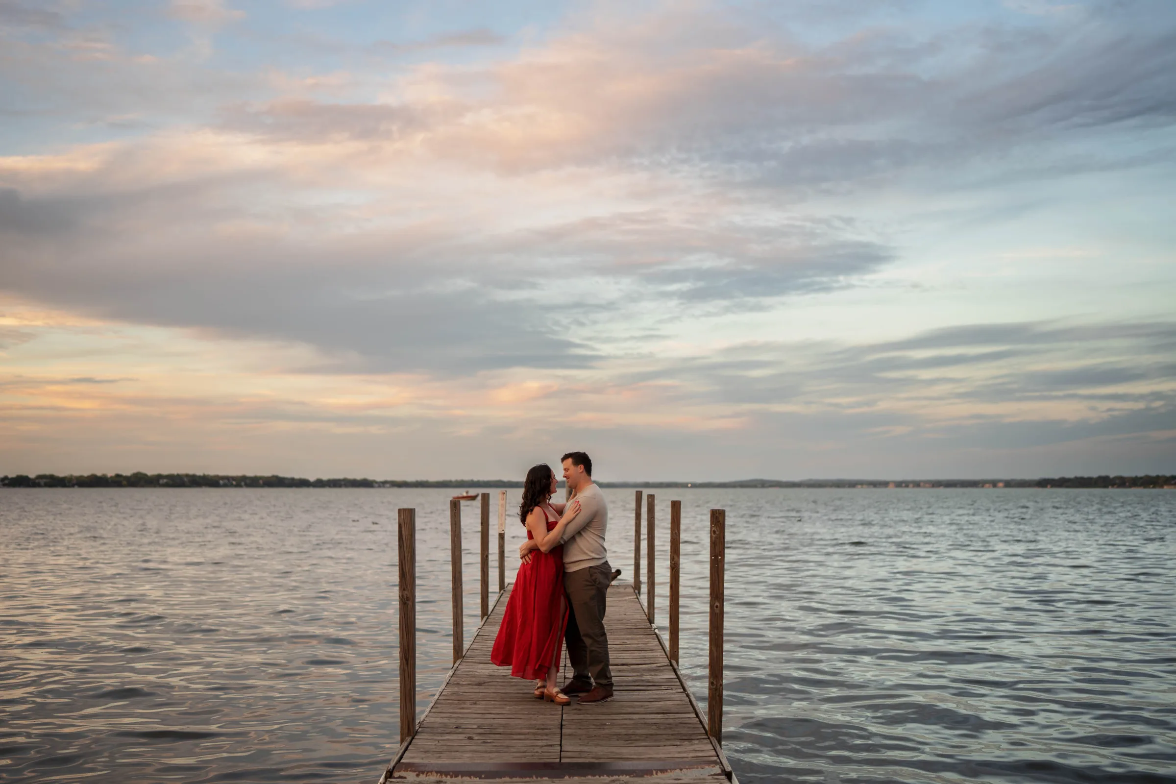 Waterfront engagement photography session in Madison, WI, by Blu Photography & Design LLC, featuring a couple embracing on a wooden lake dock at sunset under a pastel sky.