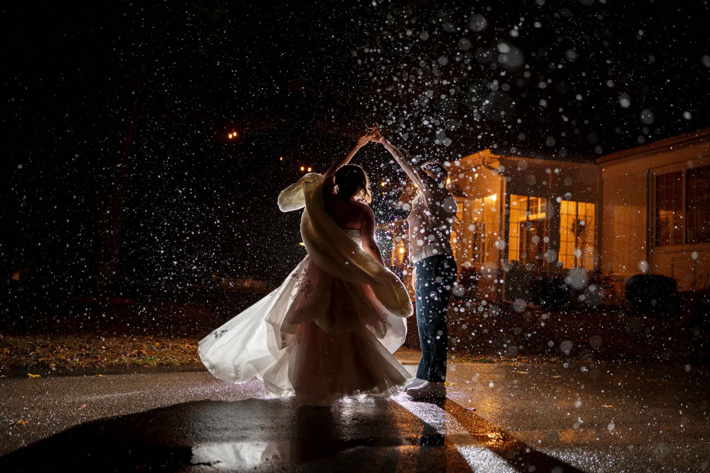 Dramatic nighttime wedding photography in Madison, WI, by Blu Photography & Design LLC, capturing a backlit couple dancing outside a venue at night as water or snow droplets create a glowing, magical bokeh effect.
