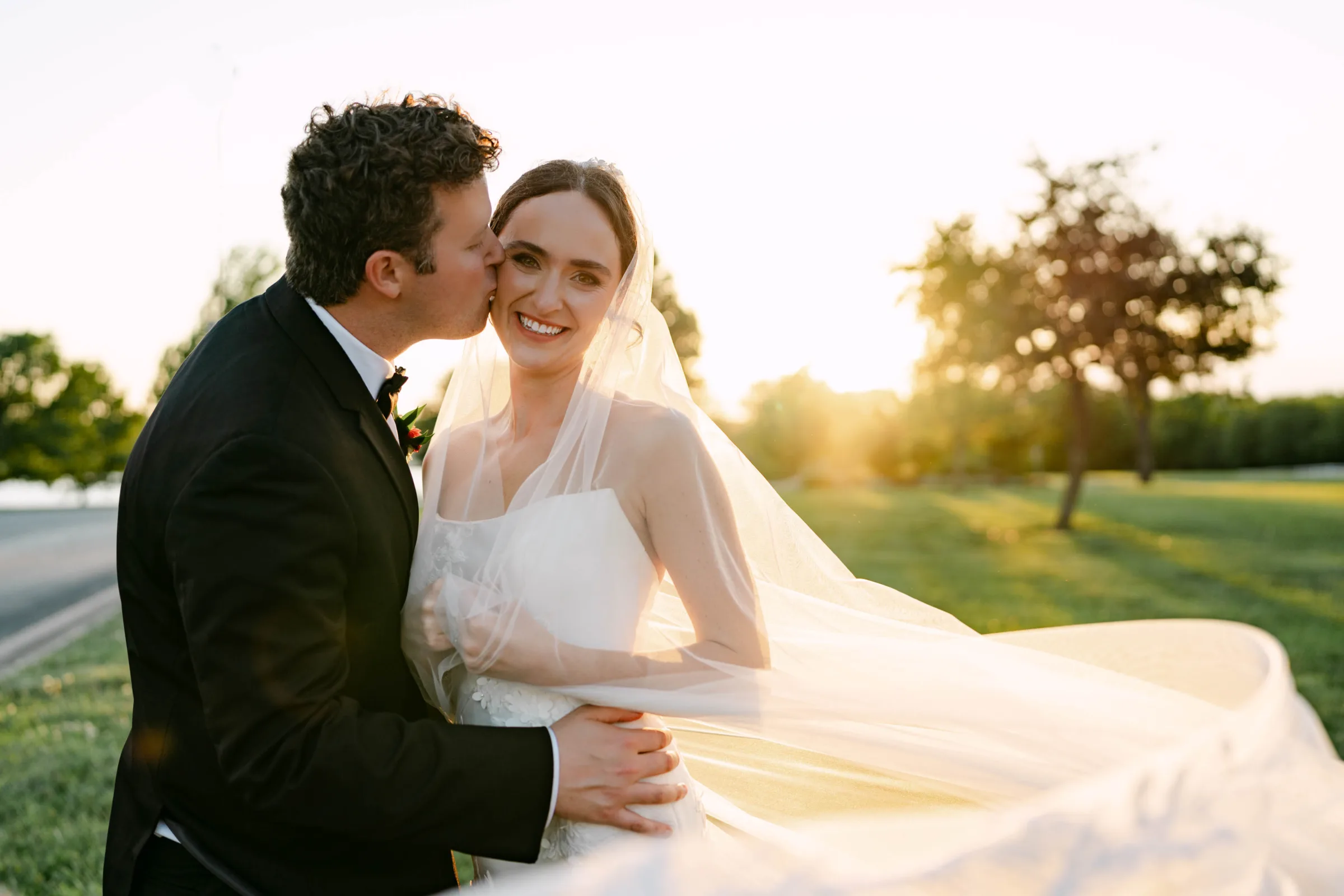 Sunset wedding portrait photography in Madison, WI, by Blu Photography & Design LLC, showing a groom in a classic tuxedo kissing a smiling bride on the cheek while her long tulle veil catches the golden hour light.