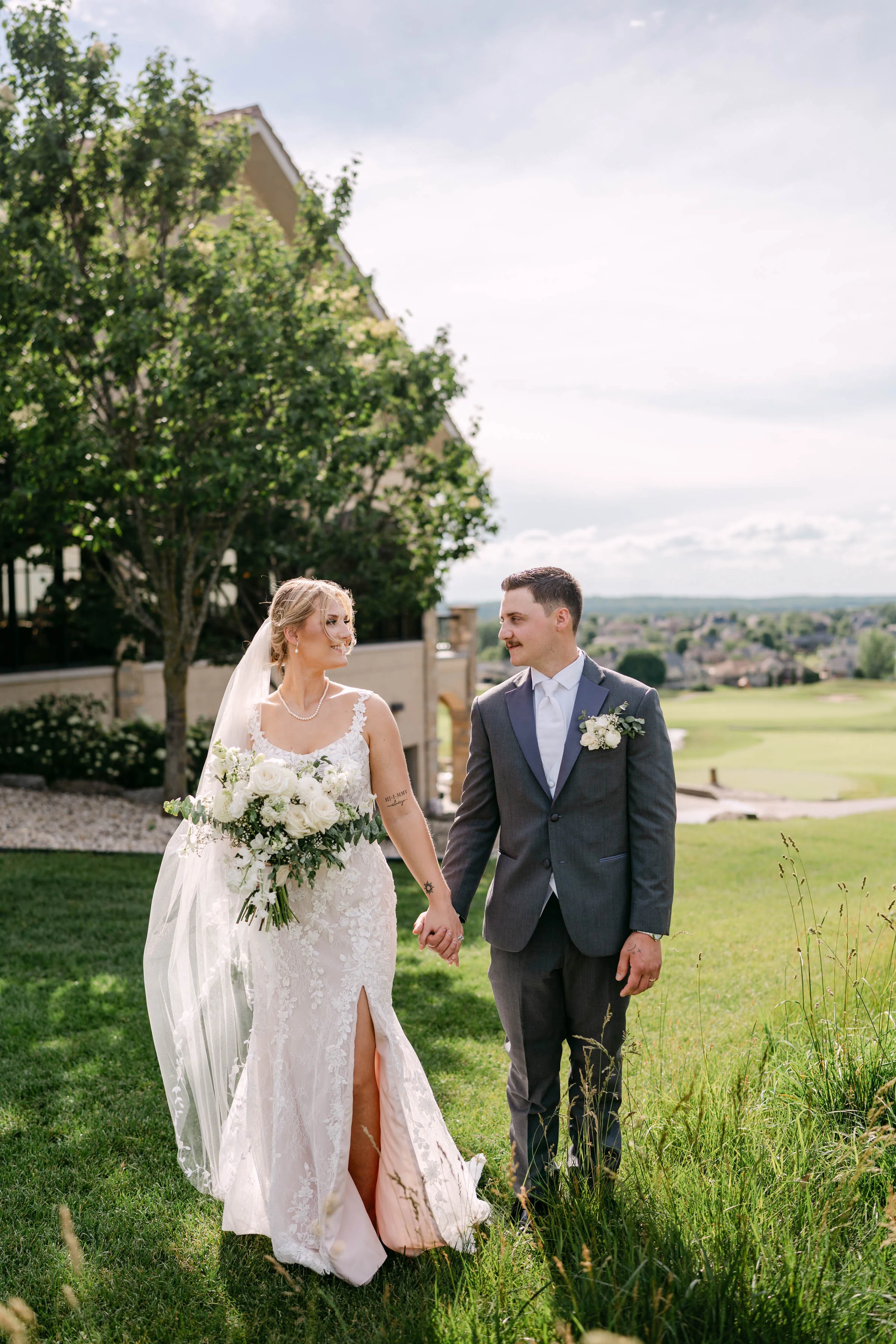 Scenic country club wedding photography in Madison, WI, by Blu Photography & Design LLC, featuring a bride in a lace dress with a high thigh slit holding hands with a groom in a grey tuxedo on a grassy hillside.