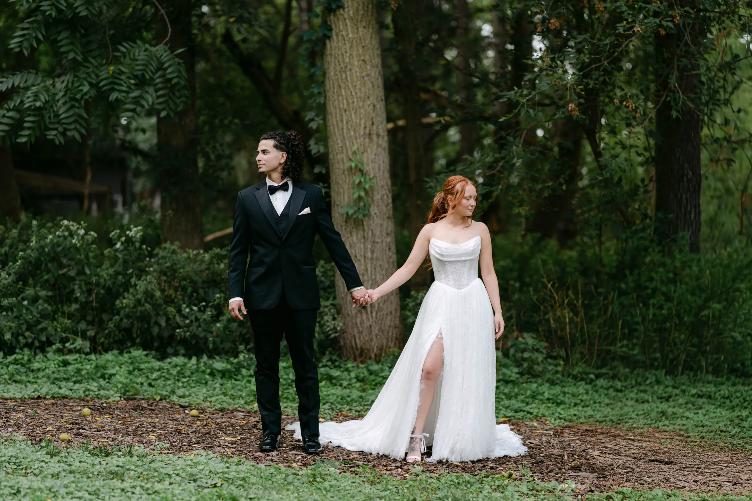 Forest wedding photography in Madison, WI, by Blu Photography & Design LLC, featuring a groom in a black tuxedo and a bride in a strapless gown with a leg slit holding hands in a lush, wooded park.