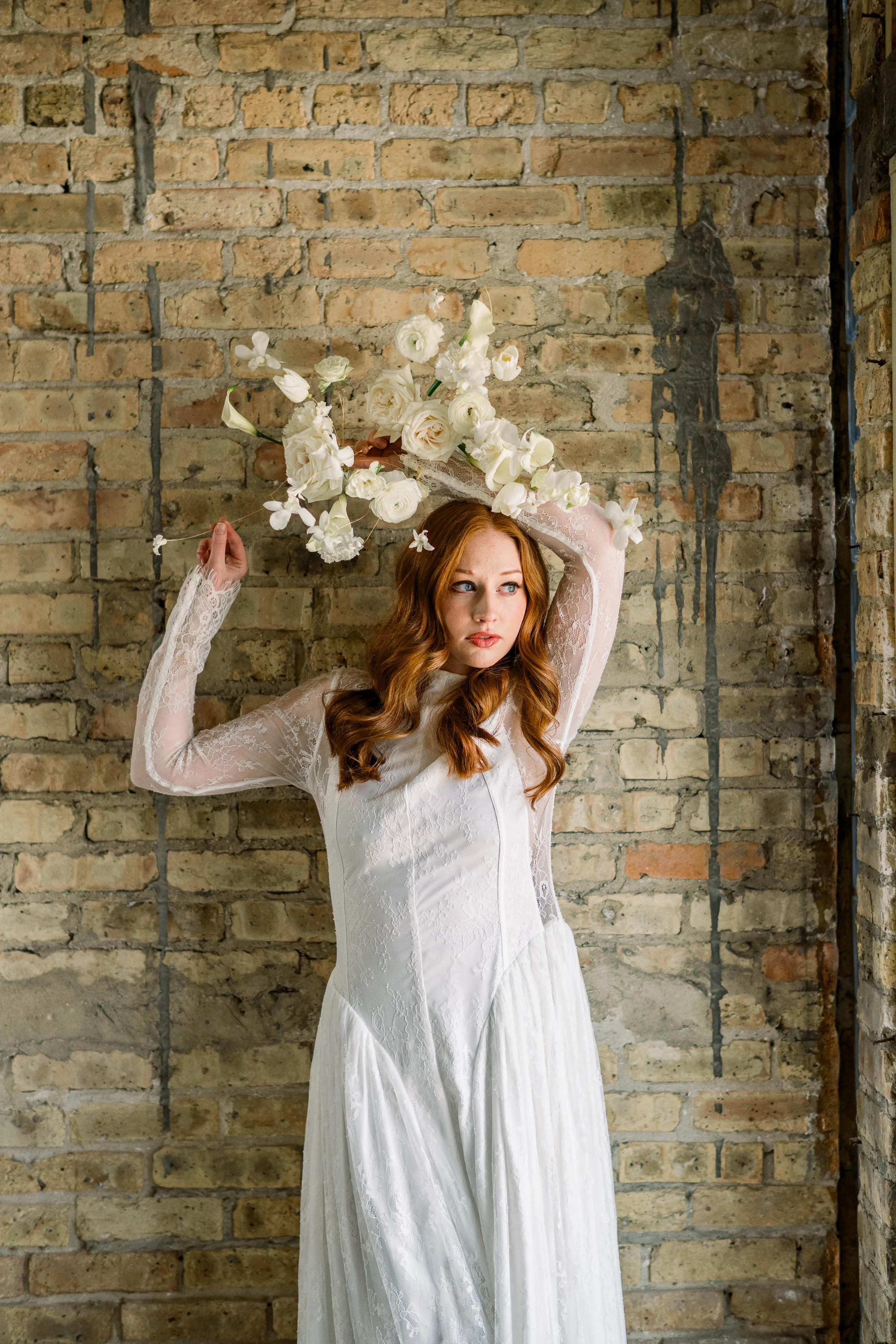 Contemporary white bridal bouquet designed by Cosmic Gardens in Cross Plains, WI, held artfully above the head of a red-haired bride in a long-sleeve lace gown against a rustic brick wall.