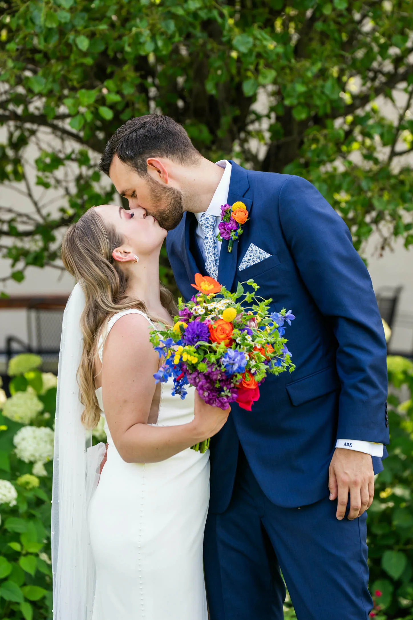 Vibrant wildflower wedding bouquet and matching boutonniere by Cosmic Gardens in Cross Plains, WI, featuring bright orange poppies, purple delphinium, and yellow billy balls held by a bride and groom kissing.
