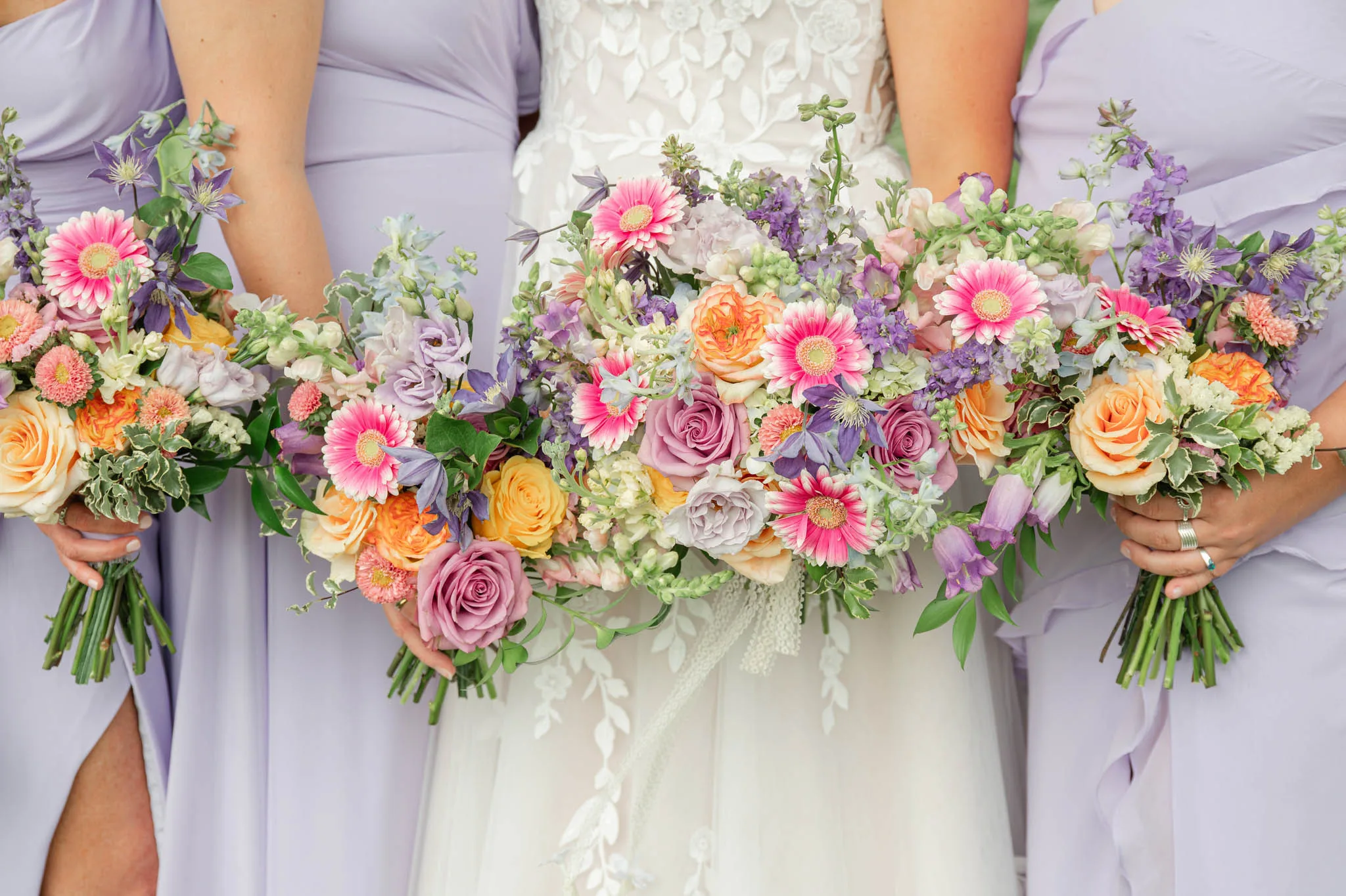 Pastel lavender, pink, and peach wedding bouquets by Cosmic Gardens in Cross Plains, WI, featuring bright pink gerbera daisies, lavender roses, and larkspur held by a bride and her bridesmaids in lilac dresses.