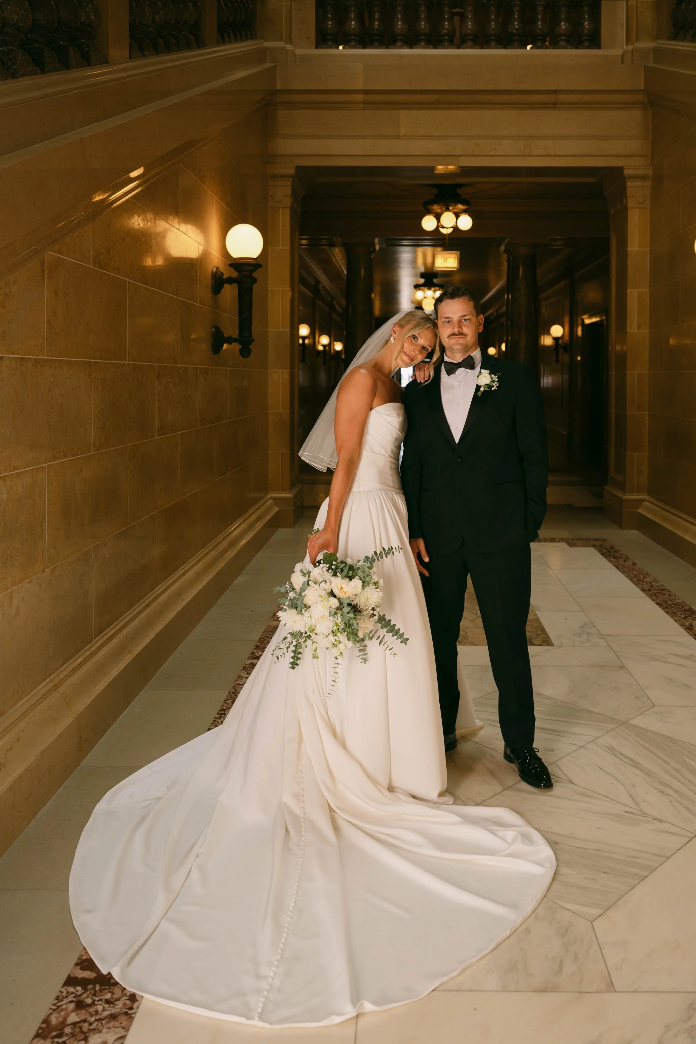 A full-length indoor wedding portrait of a bride and groom posing in a historic marble hallway, featuring a classic white bouquet and boutonniere by Sweet Pea Floral. The bride tilts her head against the groom's shoulder while holding her elegant white rose and eucalyptus bouquet over her long gown train, while the groom stands beside her in a black tuxedo with a matching white rose boutonniere.