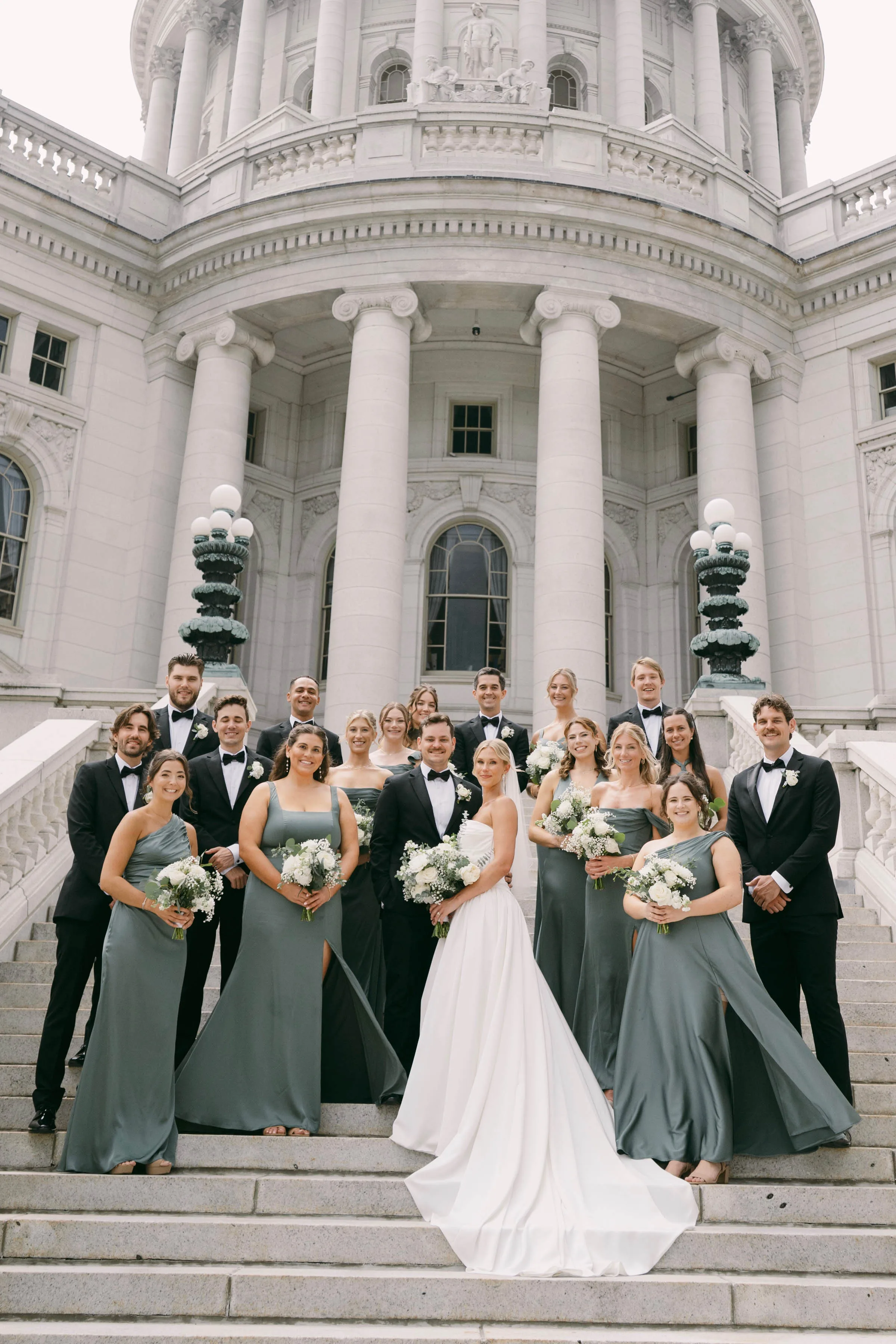 A large wedding party posing on the stone steps of the Wisconsin State Capitol building in Madison, showcasing bouquets and boutonnieres by Sweet Pea Floral. The bride holds a white rose bouquet while her bridesmaids wear sage green gowns and carry smaller coordinating white bouquets. The groom and groomsmen wear classic black tuxedos accented with elegant white rose boutonnieres.