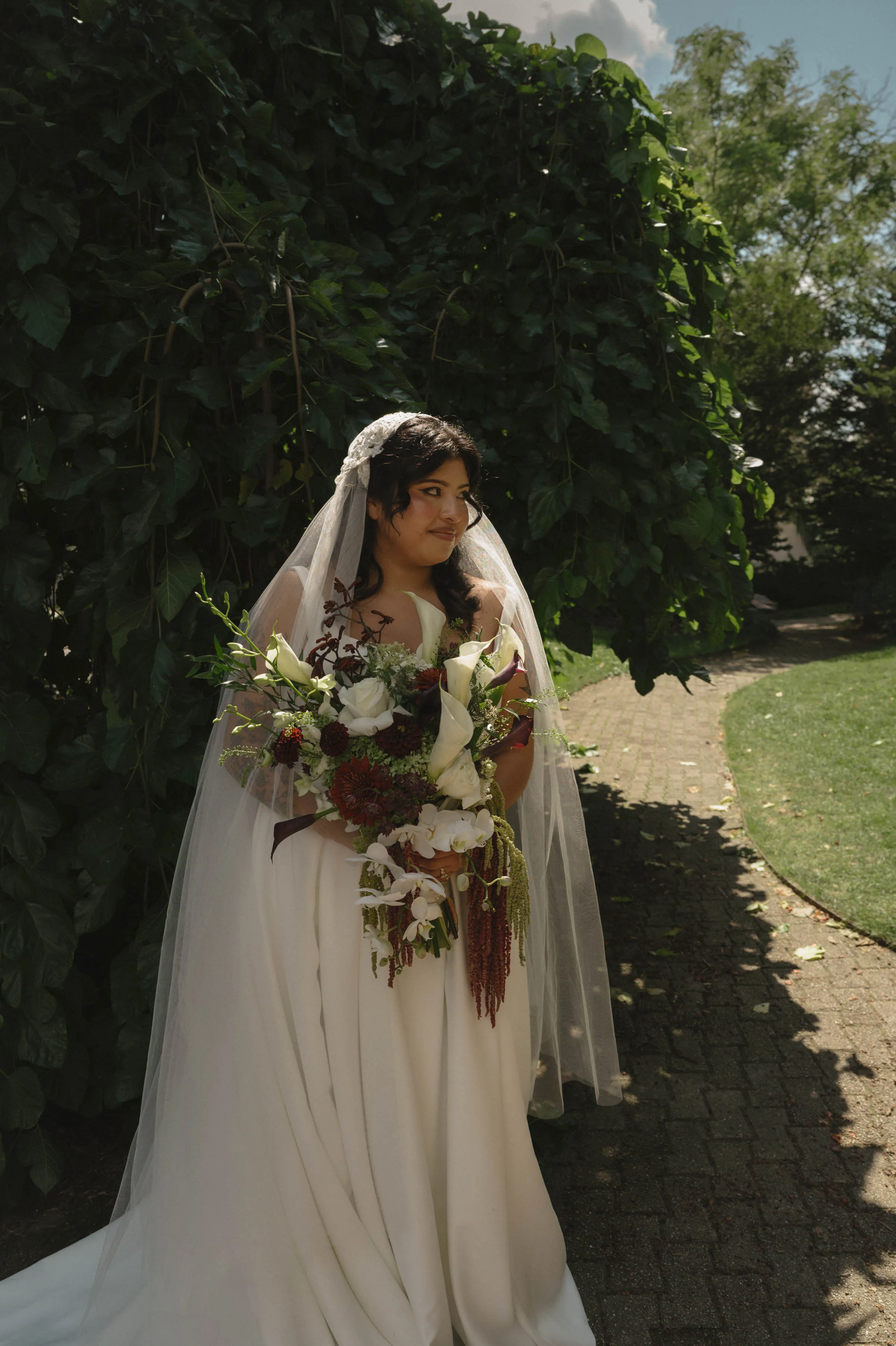 A full-length bridal portrait showcasing a dramatic cascading bouquet designed by Sweet Pea Floral. The unique arrangement features large white calla lilies, deep burgundy dahlias, white roses, and white orchids, accented with textured greenery and hanging amaranthus. The bride wears an elegant white gown with a long, sheer tulle veil and stands on a shaded brick path under a canopy of lush green leaves.