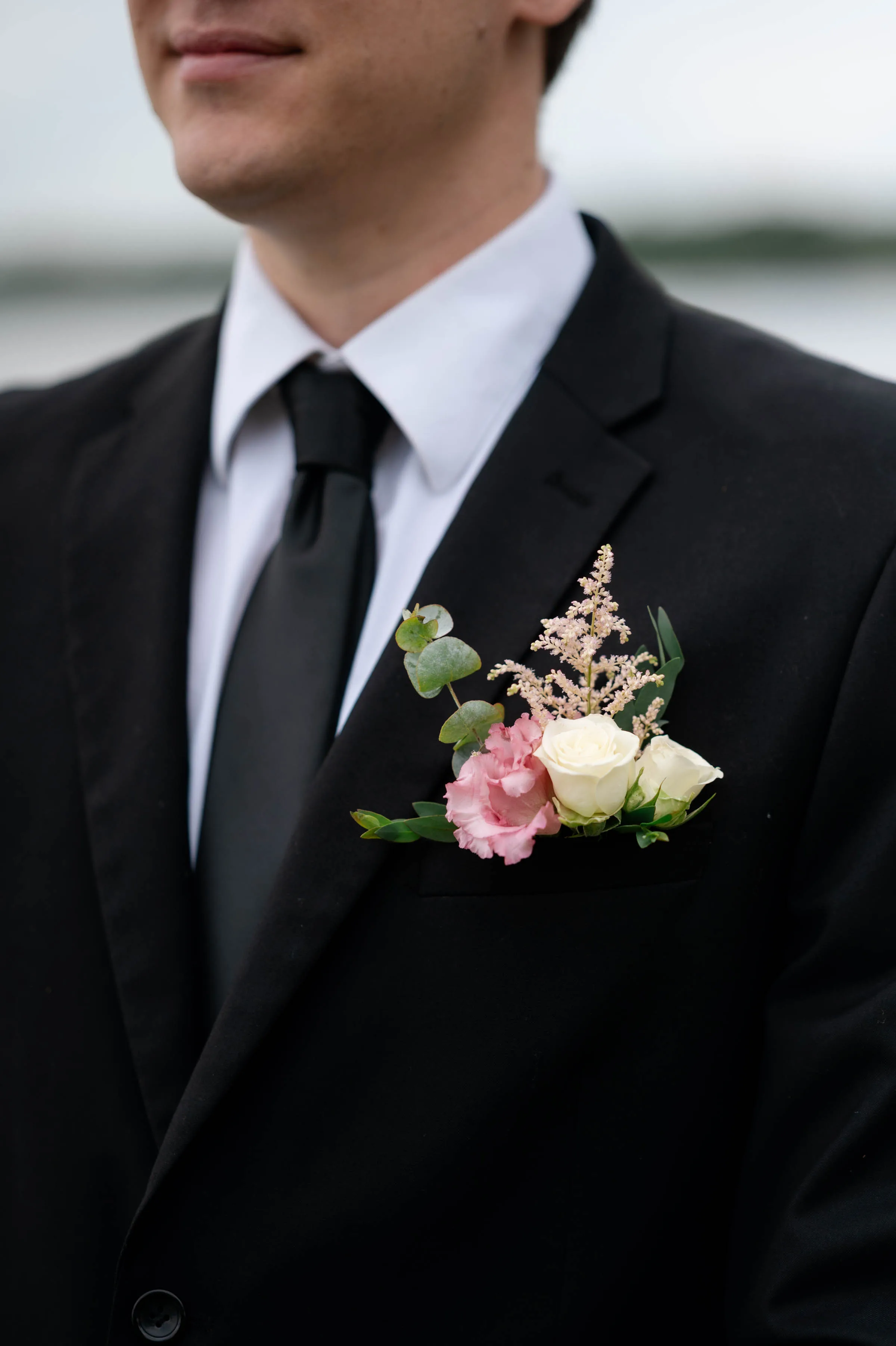 A close-up photograph of a custom wedding boutonniere designed by Sweet Pea Floral, pinned to a groom's black suit lapel. The pocket arrangement features a delicate white rose bud, a soft pink bloom, a sprig of pink astilbe, and eucalyptus leaves, contrasting against a white dress shirt and black satin tie.