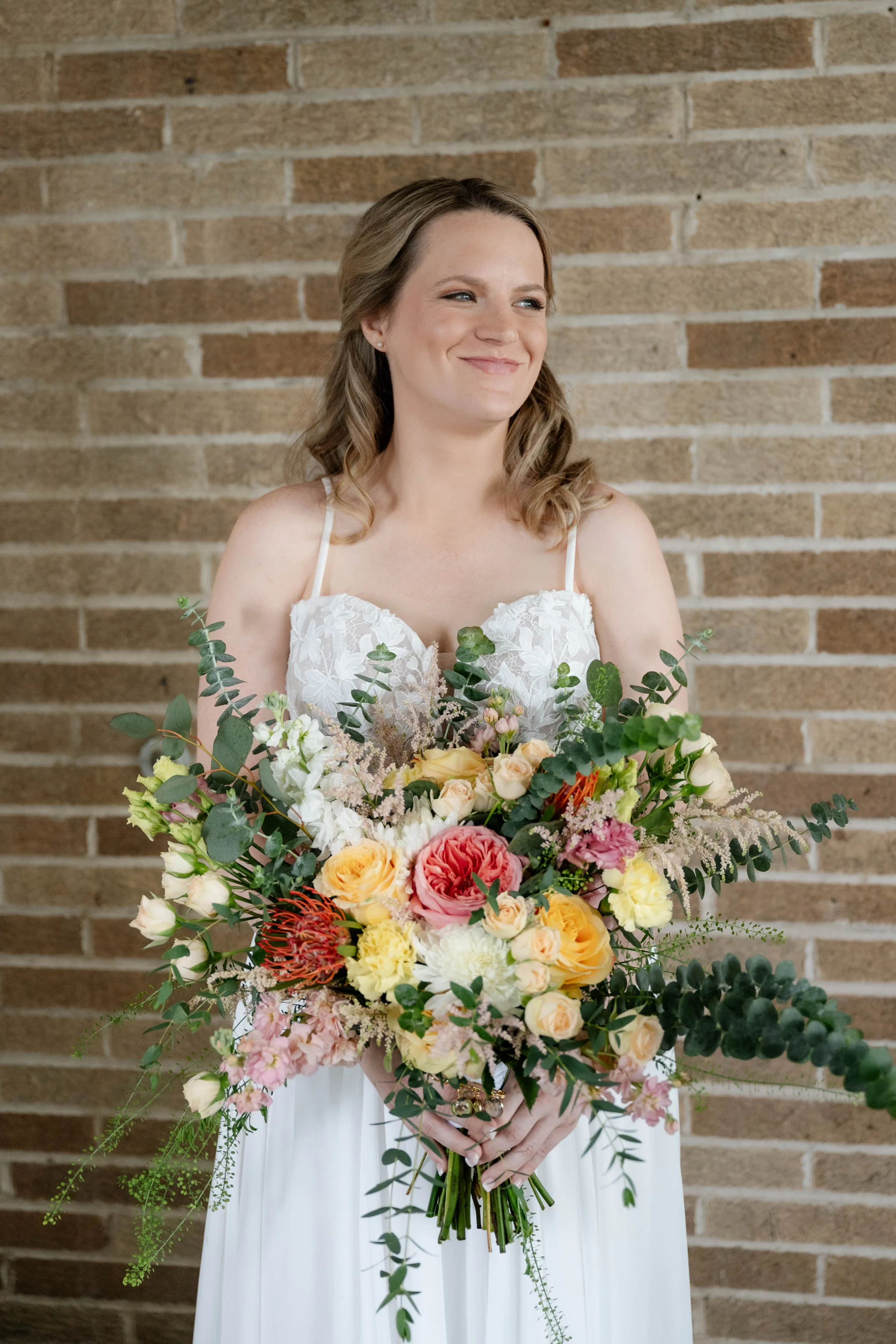 A medium-shot wedding photograph featuring an expansive, garden-style bridal bouquet designed by Sweet Pea Floral. The colorful, asymmetrical arrangement includes a prominent pink garden rose, yellow roses, orange pincushion protea, pink snapdragons, and textured sprigs of eucalyptus and pampas grass. The bride holds the bouquet in front of her white lace-bodice gown while smiling against a light brown brick wall.