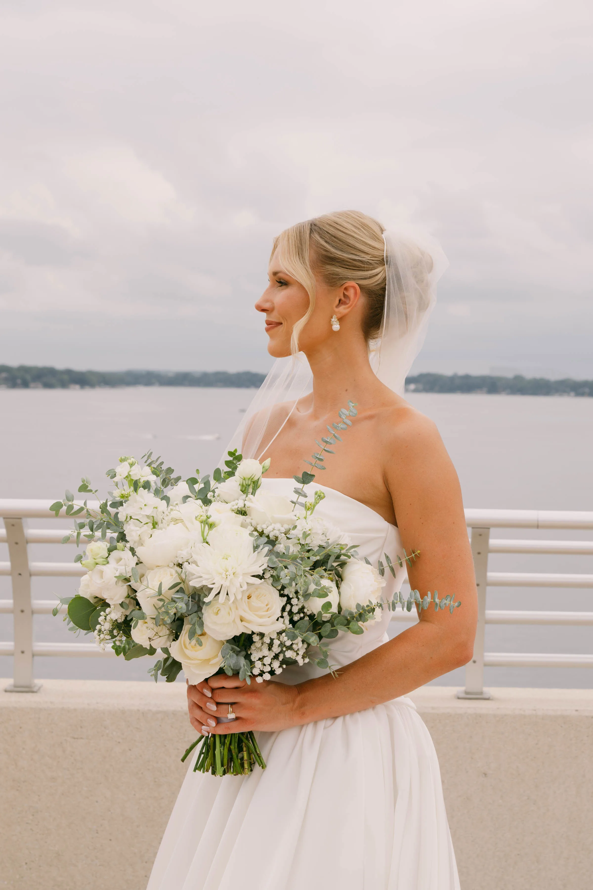 A profile wedding portrait highlighting an all-white bridal bouquet designed by Sweet Pea Floral. The textured arrangement features white roses, chrysanthemums, ranunculus, and baby's breath, accented with sprigs of silver dollar and spiral eucalyptus. The bride, wearing a strapless white gown and short veil, stands on an outdoor terrace looking out over a wide lake under an overcast sky.