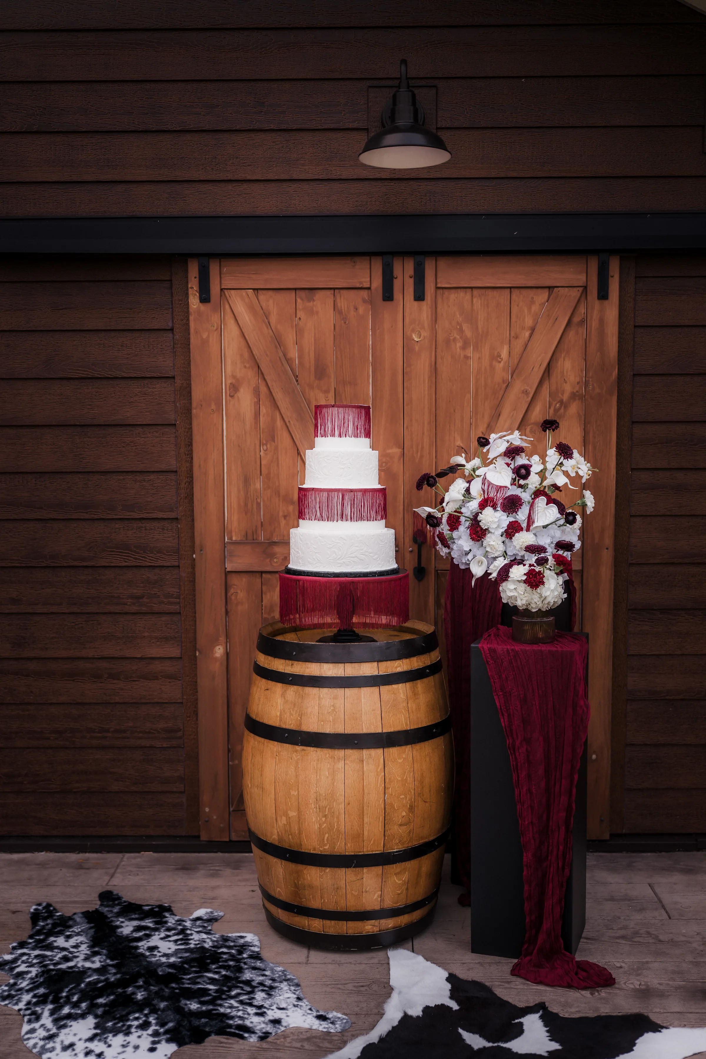 a white, four tier, textured wedding cake from Craig's Cake Shop with burgundy tassels around the second and top layers. The cake is presented on a rustic wooden barrel beside another table with decorative florals