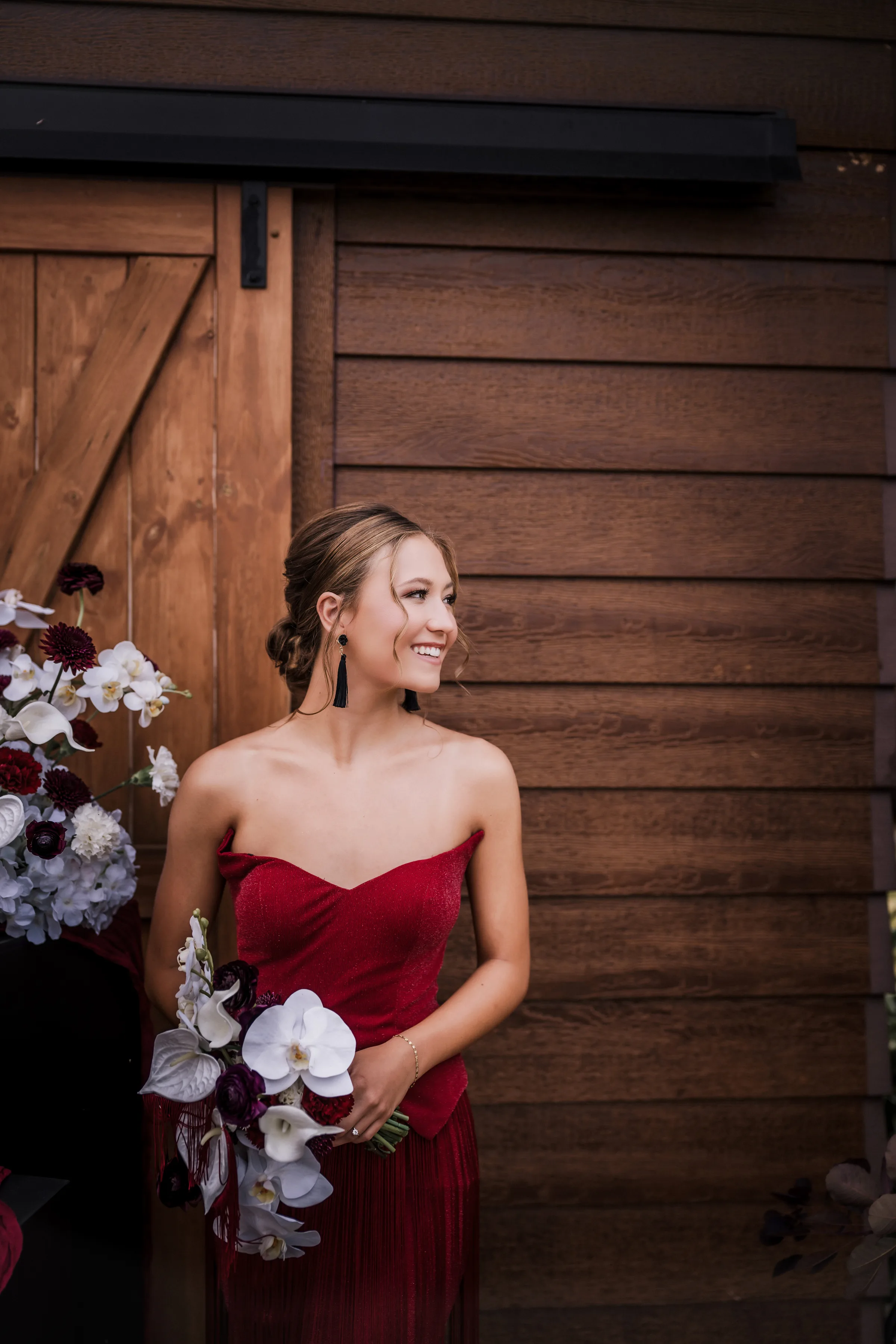 a bridesmaid wearing a sleek burgundy dress smiles in front of a rustic barn while holding a bouquet
