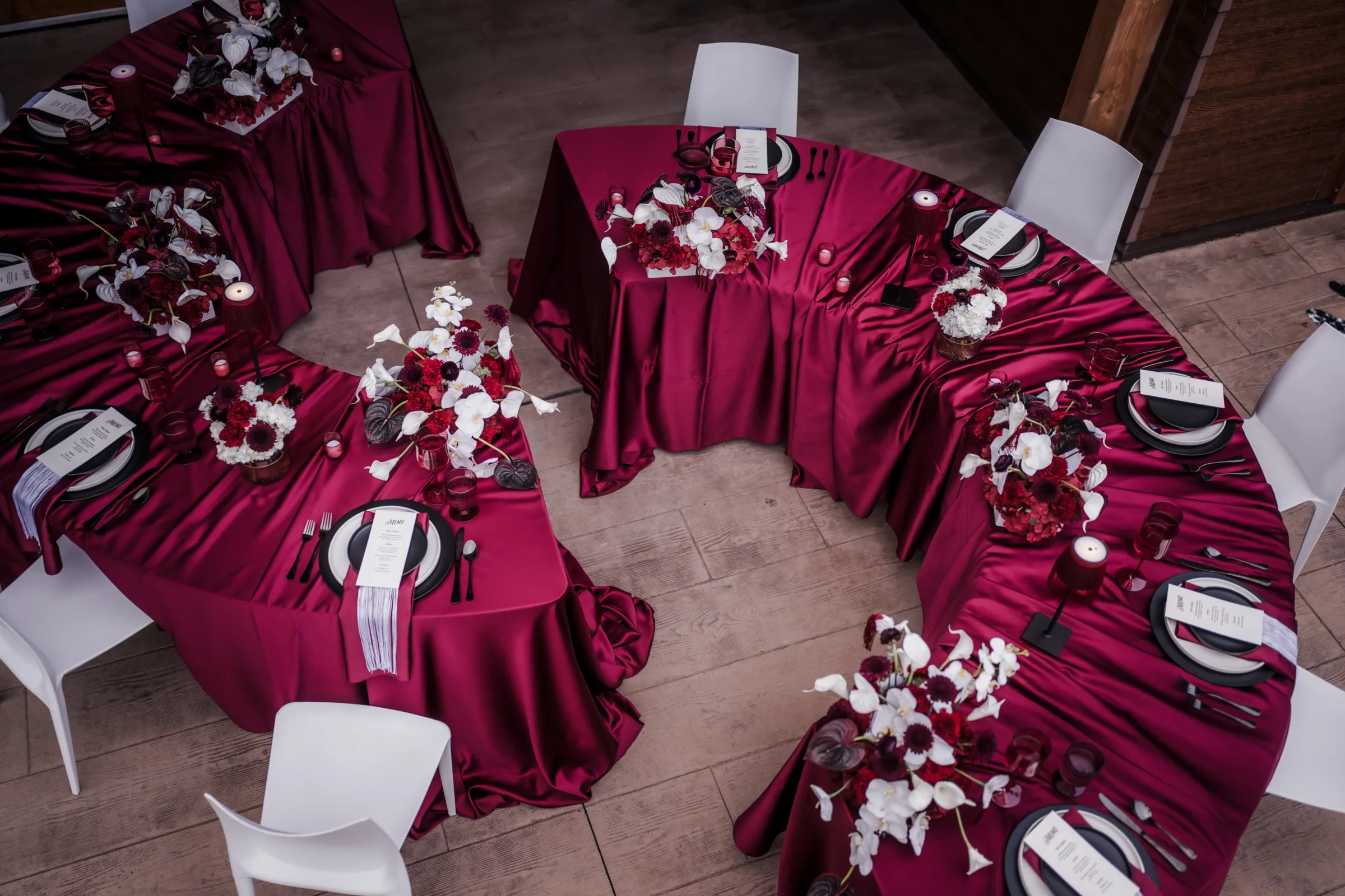 an overhead view of two half-circle wedding reception tables staggered to create an aisle between them. Both tables are draped in burgundy velvet tablecloths, and place settings consist of white chairs, layered black and white plating, burgundy linens, tinted crystal glassware, custom menus, and silverware. The styling is completed by floral centerpieces, table lanterns, and tea lights