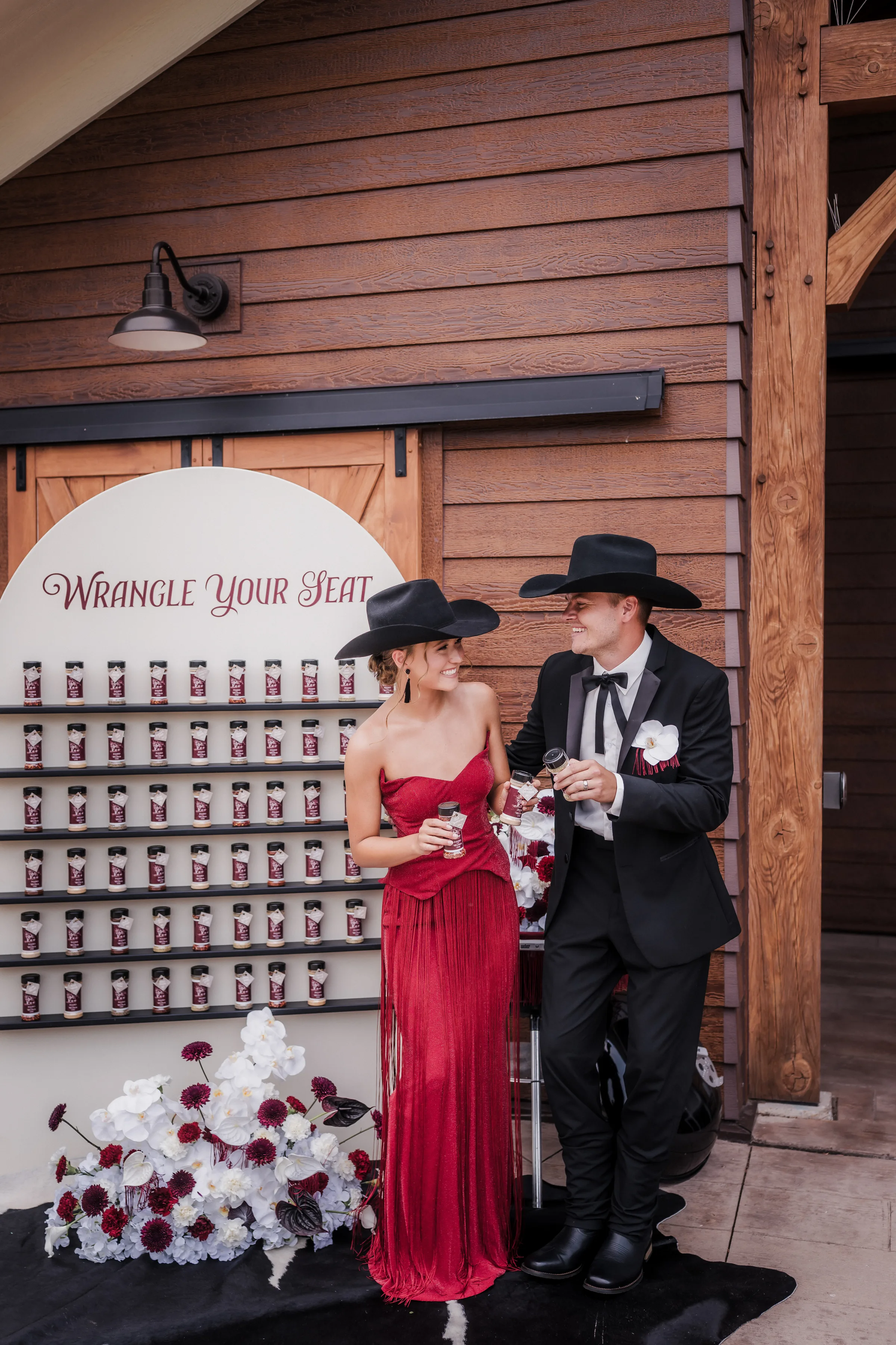 maid of honor and best man models stand beside an interactive seating chart by LK Bridal Studio with spice favors that double as escort cards. They are both wearing cowboy hats and are dressed formally for the modern cowboy inspired editorial at Barnwood Events in Madison, Wisconsin