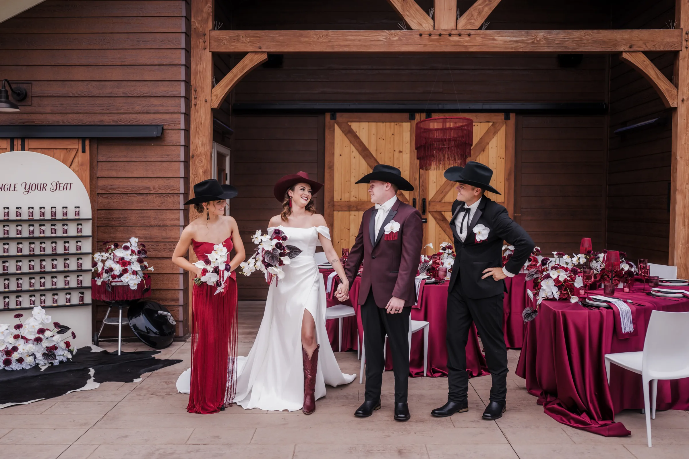 four models (bride, groom, maid of honor, best man) stand in front of a moody cowboy reception table set up at Barnwood Events. The wedding palette is white, red, burgundy, wine, and black
