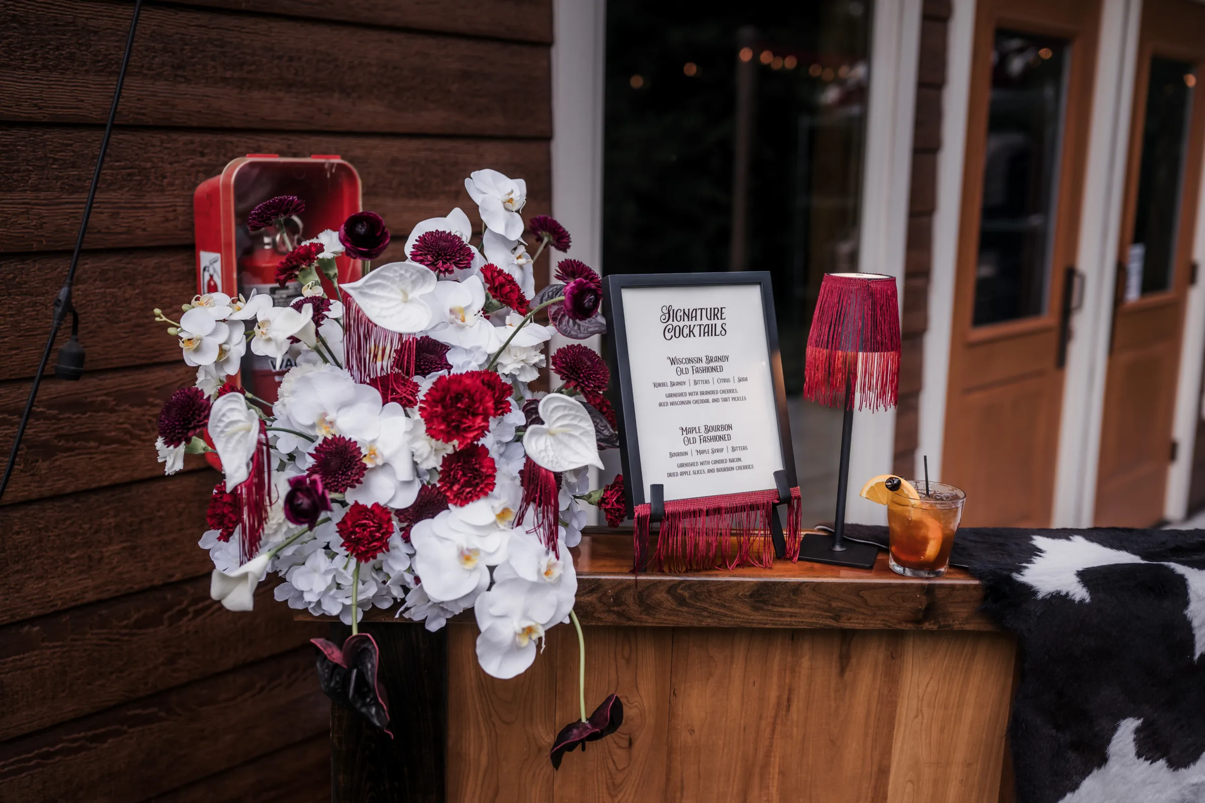 modern cowboy wedding sign bar signage from LK Bridal Studio displayed alongside white, red, and burgundy florals from Melody Rose Designs and glowing table lamps