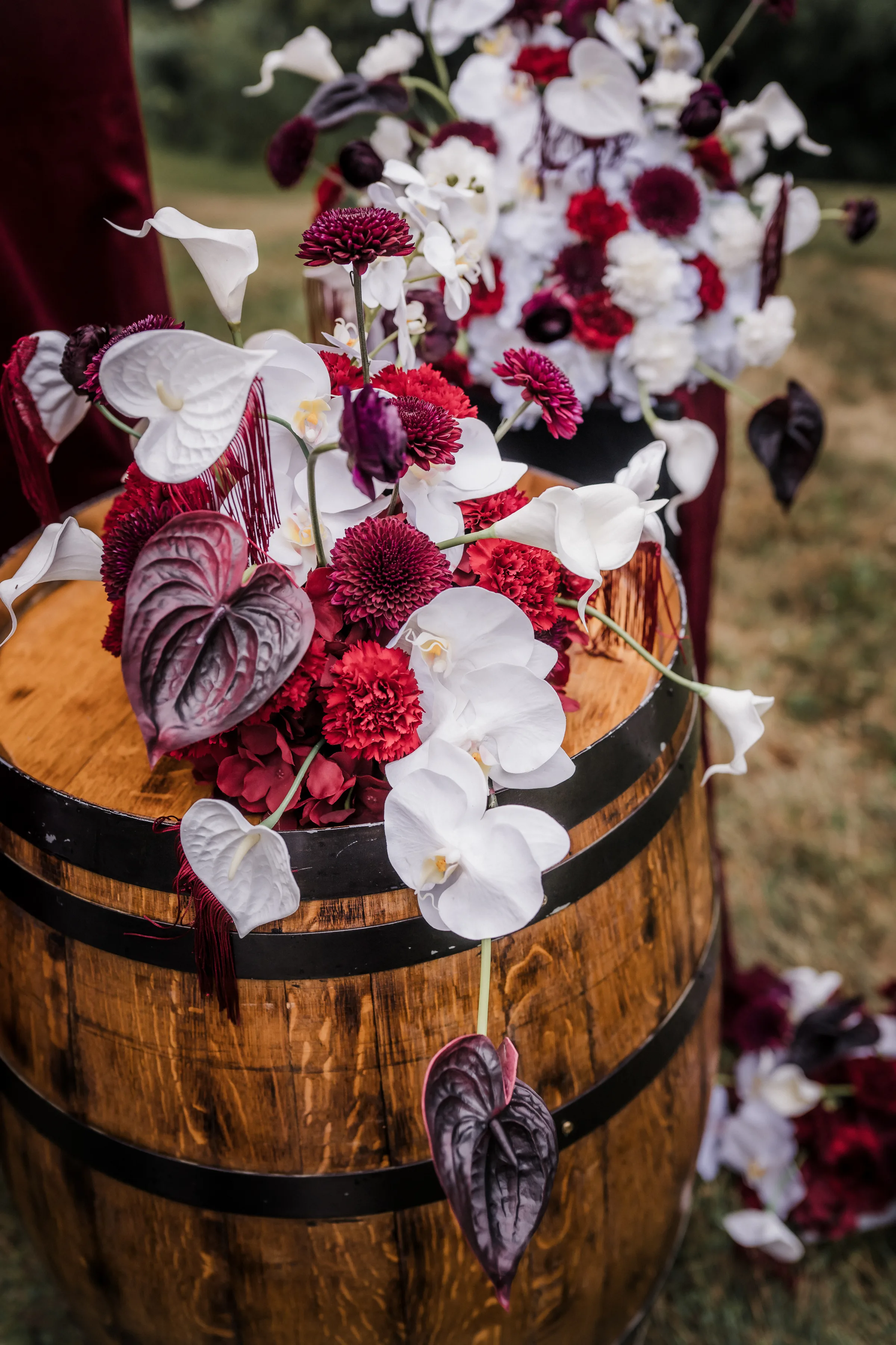 a close-up of wedding floral décor from Melody Rose Designs including white phalaenopsis orchids, white and deep burgundy anthurium, burgundy ranunculus, red dianthus, cascading burgundy amaranthus, and moody smokebush foliage.