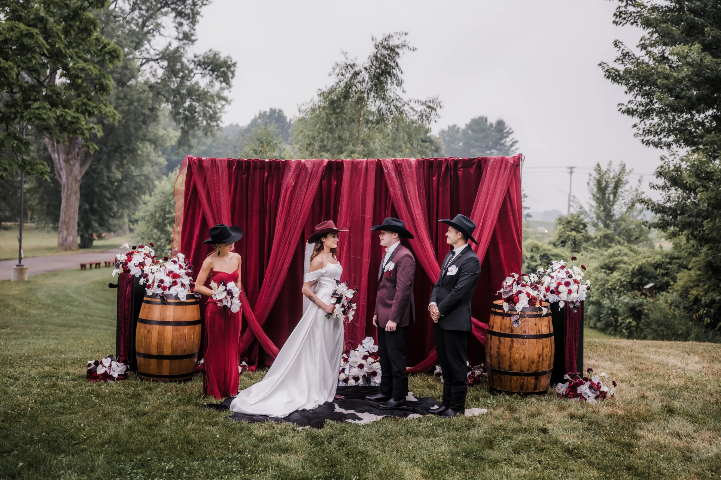 modern cowboy wedding inspiration at Barnwood Events depicting an outdoor ceremony with burgundy draping from Melody Rose Designs, wooden barrels, wooden pillars, a cow hide rug, and abundant red, white, and burgundy florals