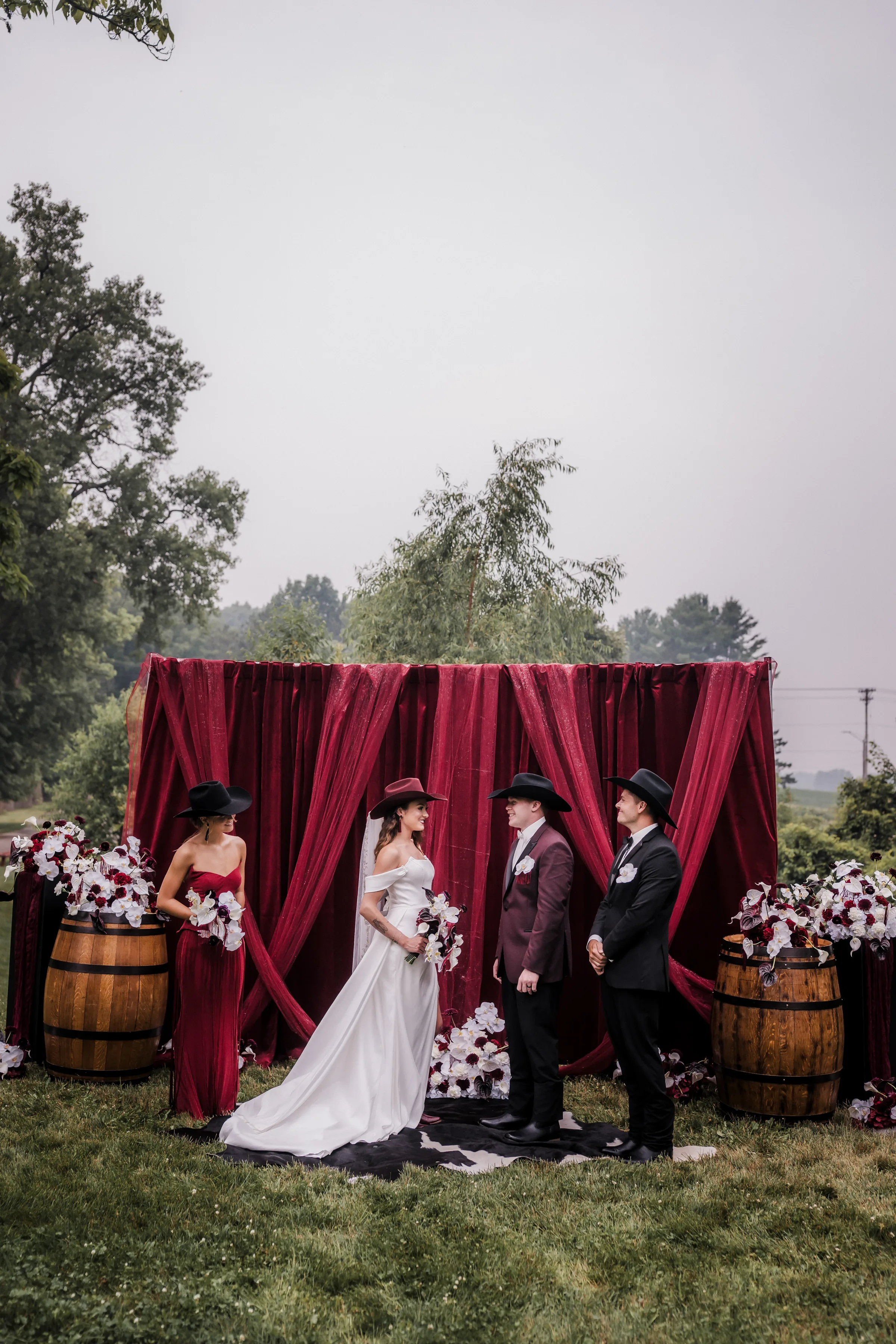 bride and groom models smile with their wedding party in front of a moody burgundy ceremony backdrop