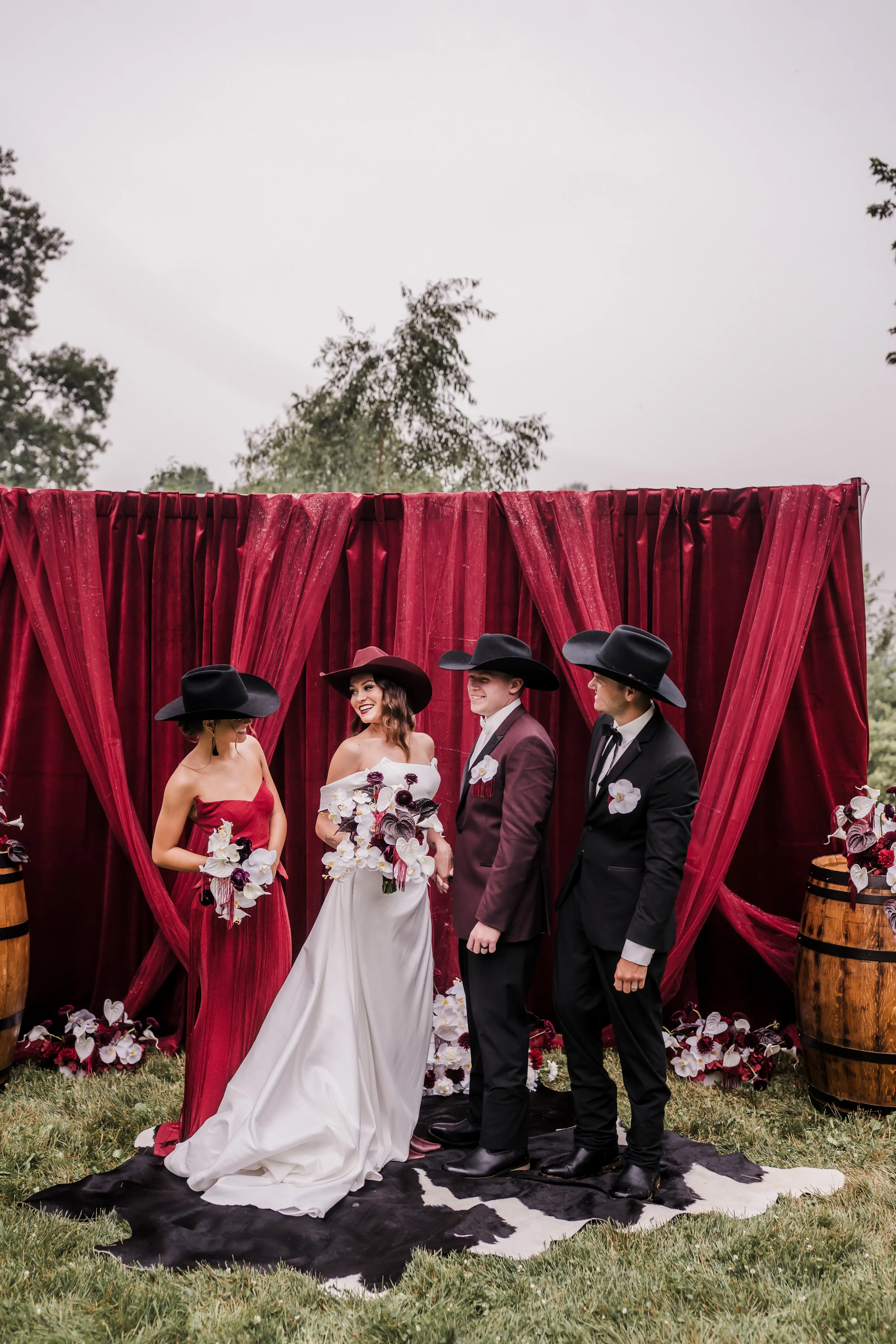a bride and groom interact with their wedding party at a moody modern cowboy wedding ceremony. All are wearing cowboy hats and dressed in either red, burgundy, black, or white