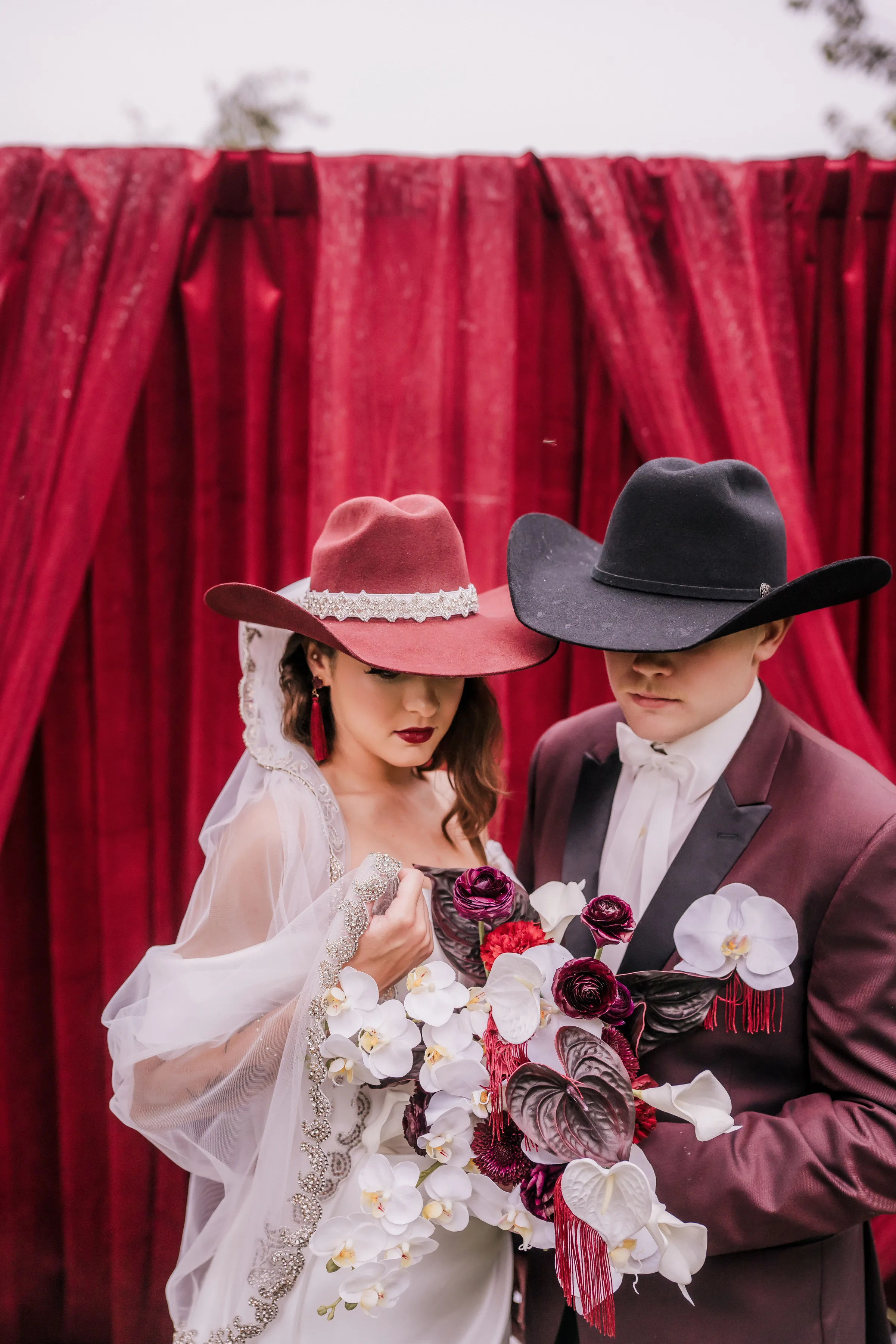 a bride and groom stand at the ceremony of a moody modern cowboy wedding. The bride is wearing a wine-colored cowboy hat with rhinestones and the groom is wearing a black cowboy hat